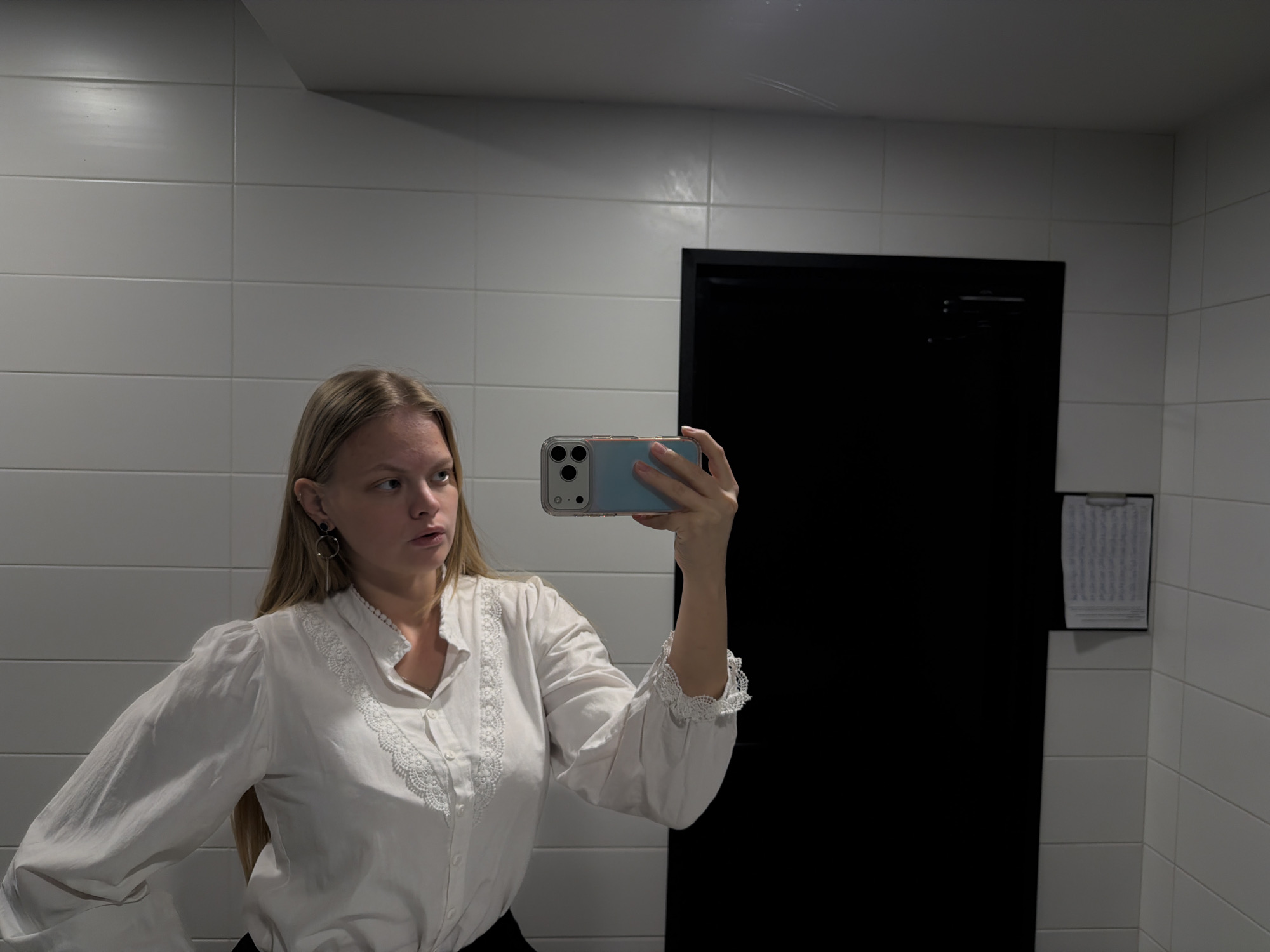 A young woman with long, light blonde hair is depicted in a mirror selfie, holding a light blue-cased smartphone with a prominent triple-camera system. She is wearing a white, long-sleeved blouse featuring intricate lace detailing along the standing collar, button placket, and gathered cuffs. A dark, circular earring with a dangling gold-toned bar is visible on her left ear. Her gaze is directed towards the phone, indicating she is looking at her reflection or composing the shot, and her expression is neutral to slightly contemplative.
The setting is an indoor room with white rectangular tiles covering the walls, separated by dark grout lines. On the right side of the image, a large, dark, rectangular panel, possibly a door or a dark cabinet, stands against the tiled wall. To the right of this dark panel, a clipboard holding a document with faint, illegible rows of text is mounted on the wall. The lighting appears to be artificial overhead light, creating subtle reflections on the tiled surfaces. The scene is located in Izhevsk, Russia.