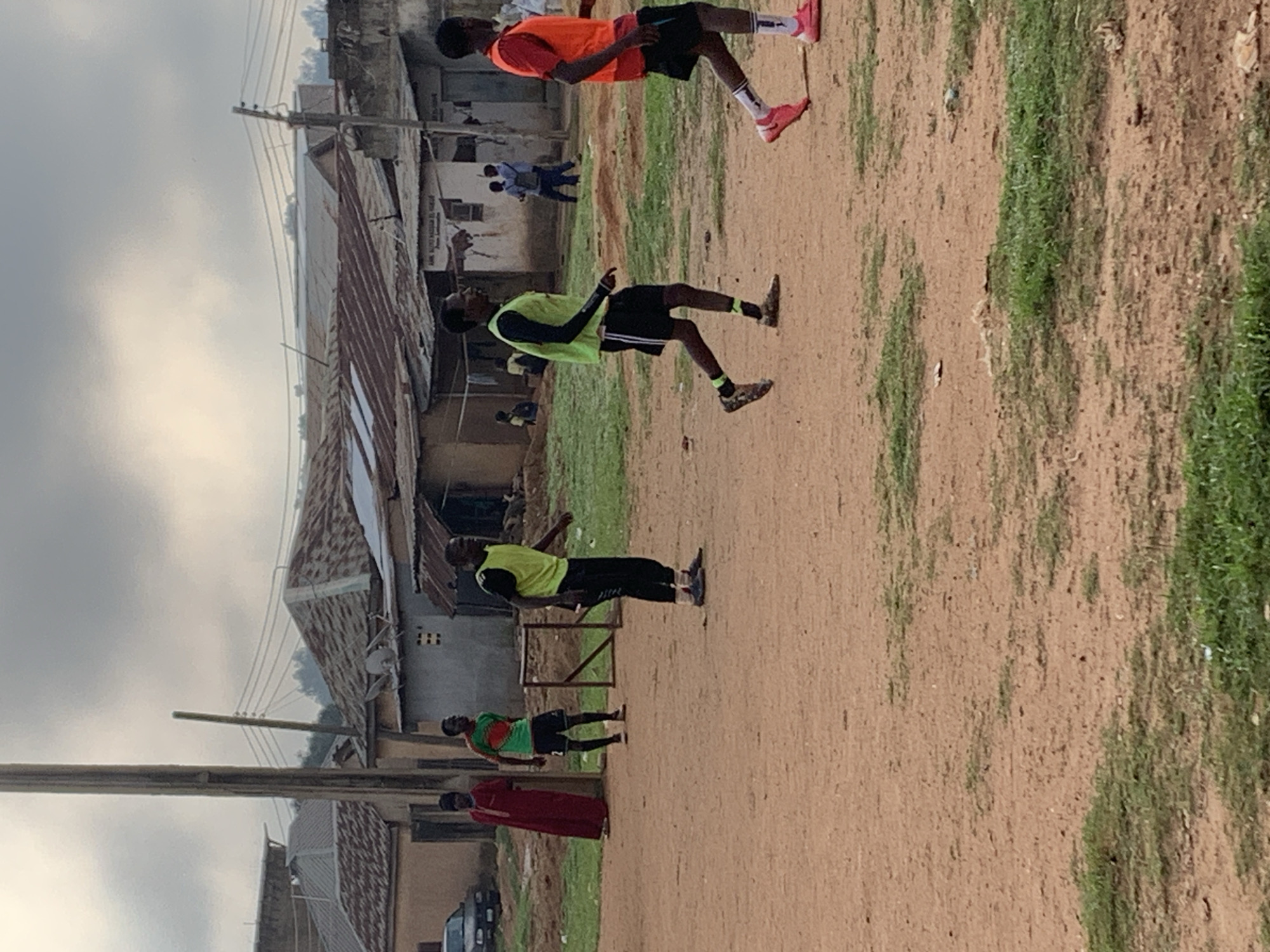 The image depicts an outdoor scene in Bukuru, Nigeria, featuring a group of young men engaged in what appears to be a casual soccer game on a dirt field. The environment suggests a semi-urban or rural setting, with a mix of simple structures in the background, some with corrugated metal roofs. A wooden soccer goal is visible on the left side of the frame.
Several individuals are in motion. In the foreground and mid-ground, three young men are prominently featured. One is wearing a bright orange vest over a red shirt and black shorts, actively running. To his left, another young man in a lime green vest over black shorts is also running. Further back, a young man in a green and red patterned shirt with black shorts is standing near the goal. To the far left, a person in a long red garment is standing. In the background, other figures are sparsely visible, including two individuals standing near a building and another person sitting.
The ground is predominantly dusty earth with patches of sparse green grass. The sky is overcast with grey clouds. Power lines and poles are present, indicating the presence of infrastructure. The overall impression is a snapshot of everyday life and recreation in a Nigerian community.