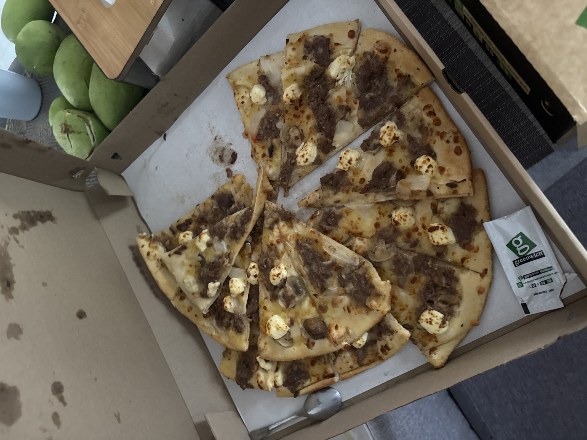 The image captures an overhead view of an open cardboard pizza box containing multiple slices of pizza, positioned in an indoor setting. The pizza features a topping of ground meat, sliced white onions, small clusters of white cheese, and some visible mushrooms on a light-colored crust. Grease stains are present on the interior surface of the pizza box lid. A silver spoon is resting within the pizza box on the left side. To the right of the pizza box, approximately five to six green, oval-shaped fruits, consistent with unripe mangoes, are arranged on a wooden or laminate surface. One of these fruits displays a noticeable crack or cut. A light-colored mug is partially visible behind the fruits. A white sachet with the "Greenwich" brand logo in green, along with delivery website and phone number details, is visible inside the pizza box at the bottom right. A gray upholstered surface is partially visible on the far left, while a dark gray textured mat is positioned beneath the right side of the pizza box. The scene depicts a casual meal in a domestic environment, consistent with a residence in Davao City, Philippines, given the identification of the Greenwich brand, a prominent Philippine fast-food chain with a presence in the city.