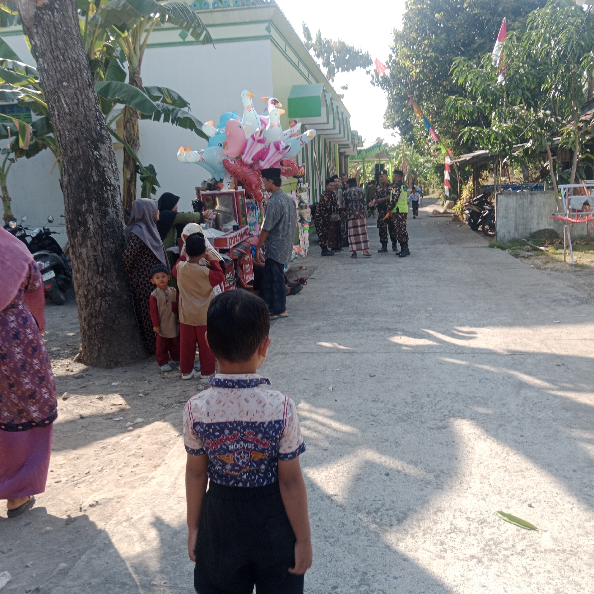 In Tegowanu Kulon, Indonesia, on a sunny day, a street scene unfolds with several individuals. In the foreground, a young boy is facing away from the camera, wearing a patterned short-sleeved shirt and dark pants. To his left, two other children, one in a tan shirt and red pants, and another in a tan top and red pants, stand near a vendor stall. The stall displays various balloons and other items. Several adults, some wearing hijabs, are gathered around the stall. Further down the street, a group of men, some in uniform, are standing together. The background features a building with a green and white facade, banana trees, and other foliage. The ground is a paved street, with a dirt area on the left.