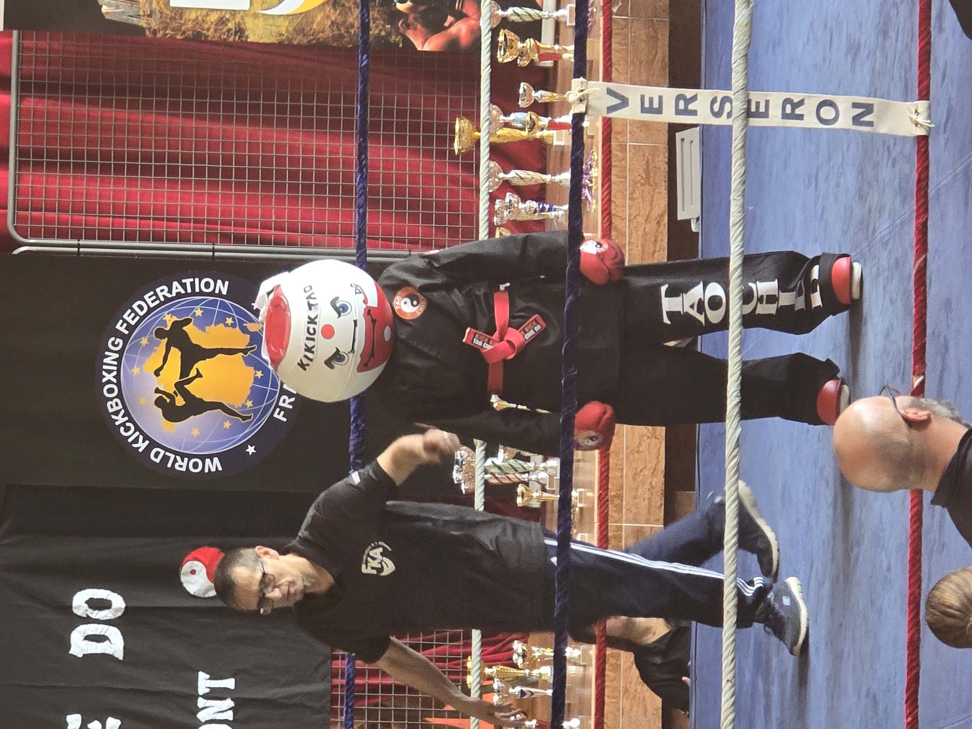 The image shows a martial arts event in Berlaimont, France. A banner displaying "WORLD KICKBOXING FEDERATION" is visible. A person in a black uniform with a red belt stands in what appears to be a boxing ring. They are wearing protective gear and a helmet with the text "KIKICK TAO". In the foreground, a person wearing glasses and a black shirt with white stripes on the pants seems to be instructing or demonstrating a technique. Multiple trophies are displayed in the background. The banner above the ring reads "VERSERON."