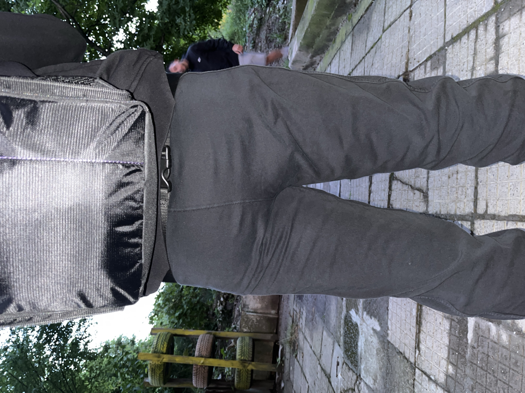 This outdoor image, captured from a low, upward-angled perspective, focuses on the lower body of a person. The individual wears dark grey or black trousers and a black, textured bag or backpack with a metallic buckle on their left side.
In the mid-ground, a pathway of light-colored, square-patterned paving stones, showing some moss and wear, stretches across the frame. Further in the background, another person is partially visible, appearing to be lying or reclining on the ground, dressed in a dark top and light shorts.
The setting is characterized by lush green foliage and trees that dominate the background and edges, indicating a natural or semi-natural environment such as a park or garden. To the right, a wooden structure adorned with several tires is visible, possibly an obstacle or a decorative feature. The natural lighting suggests it is daytime, though no harsh shadows are present. No text is discernible in the image. The scene is located in Shumen, Bulgaria.