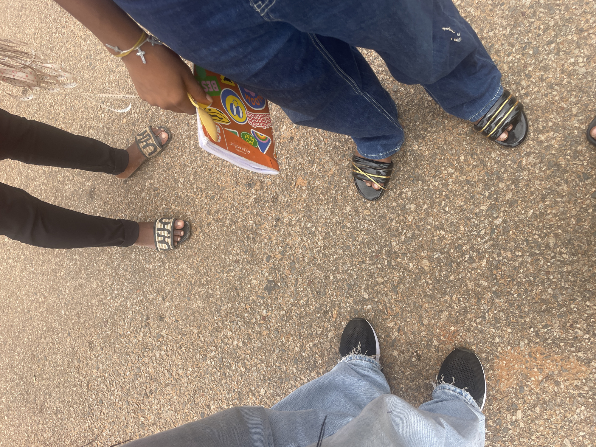In the city of Lokoja, Nigeria, four individuals are gathered on a paved surface. Their lower bodies and feet are visible, suggesting a casual outdoor setting. One person wears black pants and slides with a "CHIC" logo. Another person is dressed in dark blue jeans and black slides adorned with gold straps. A third individual is in light blue jeans, wearing black sneakers. The fourth person is also in light blue jeans, sporting black sneakers. One of the individuals is holding a colorful pamphlet or booklet. Additionally, a measuring tape is draped over the hand of the person holding the booklet. The ground appears to be asphalt or a similar paved material, with scattered pebbles and some reddish-brown stains, possibly dirt or rust. The overall scene is a snapshot of a group of people in a seemingly everyday urban environment in Lokoja.