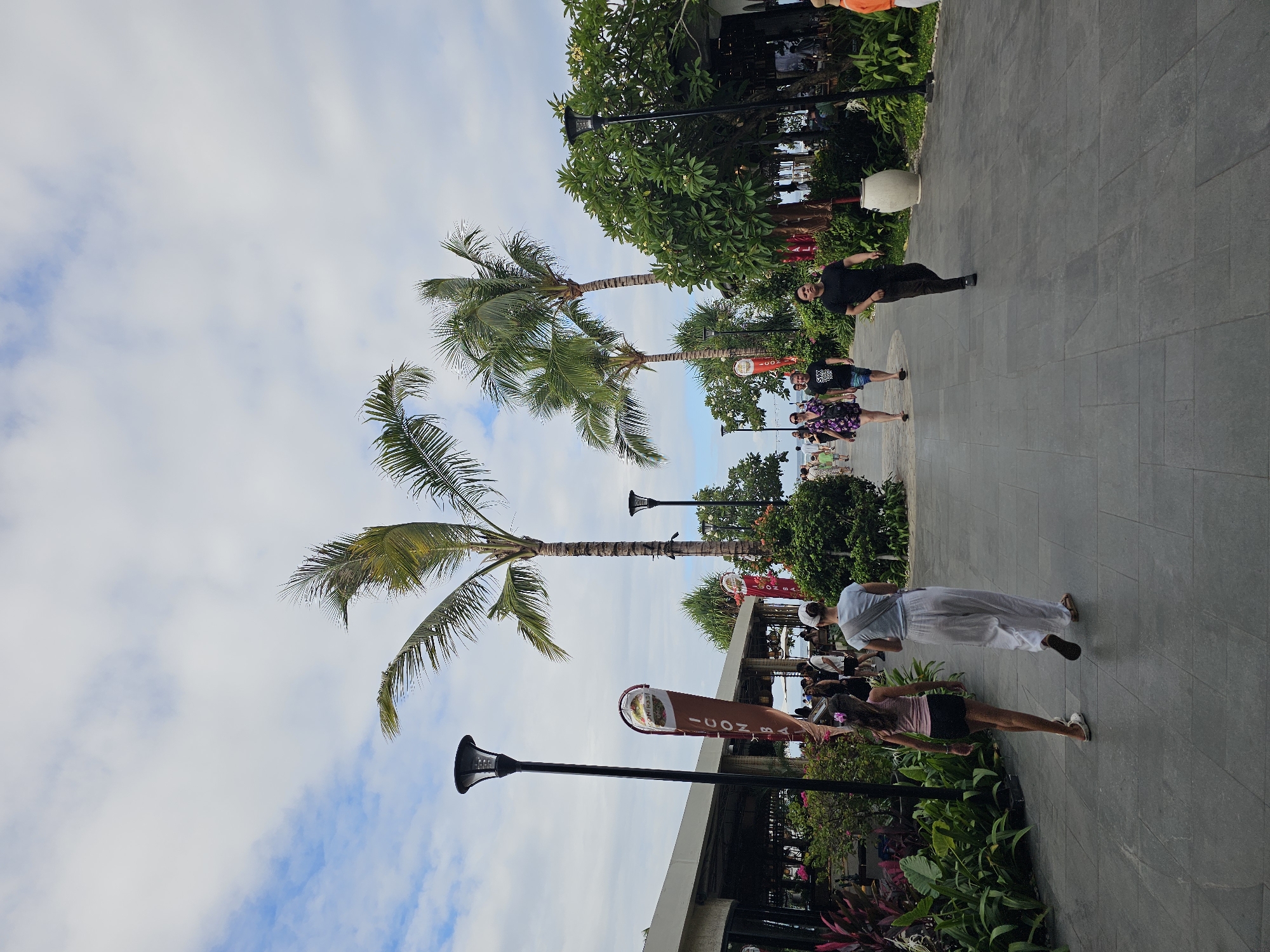 In Denpasar, Indonesia, an outdoor walkway is lined with palm trees and lush greenery. The walkway is paved with dark grey tiles. A light-colored building with an overhang is visible on the left, appearing to be a restaurant or shops. Several people are walking along the path. In the foreground, a young girl with long, dark hair and a pink tank top and black shorts walks ahead of a woman wearing a white t-shirt and loose white pants, both facing away from the camera. Further down the path, several other individuals are seen walking in both directions. Tall, black lampposts are positioned along the walkway. Red and white banners with text are hung on poles. The sky is partly cloudy with a light blue hue.