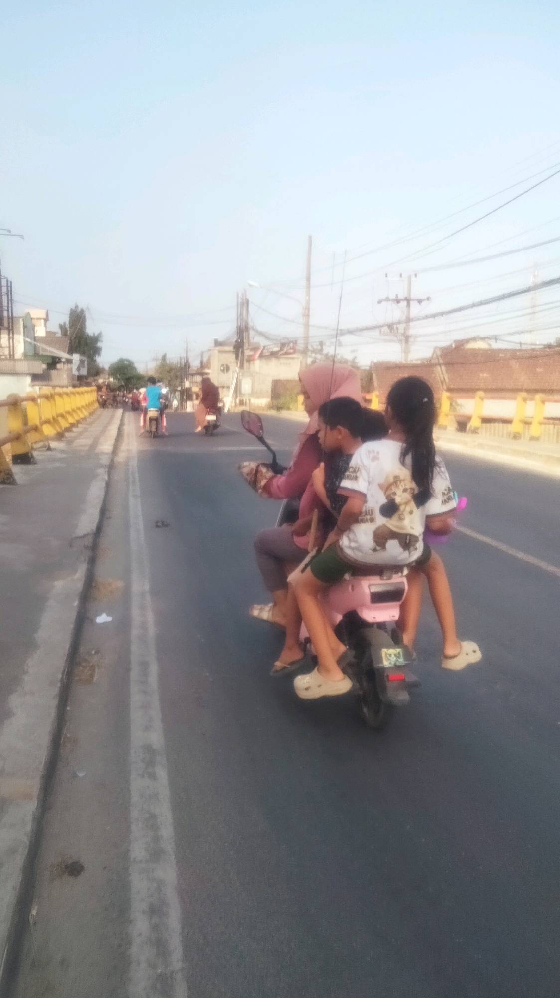 A family of three rides a pink scooter on a paved road in Palang, Indonesia. The scooter is driven by a woman wearing a pink headscarf and a dark pink top, with a younger child in a dark shirt sitting in front of her. A girl in a white t-shirt with a cartoon graphic sits behind the younger child. All three are wearing sandals. The scooter has a license plate on the back. The road is lined with a yellow-railed bridge on the left and buildings on the right, with utility poles and wires visible in the background. The sky is clear and pale blue.