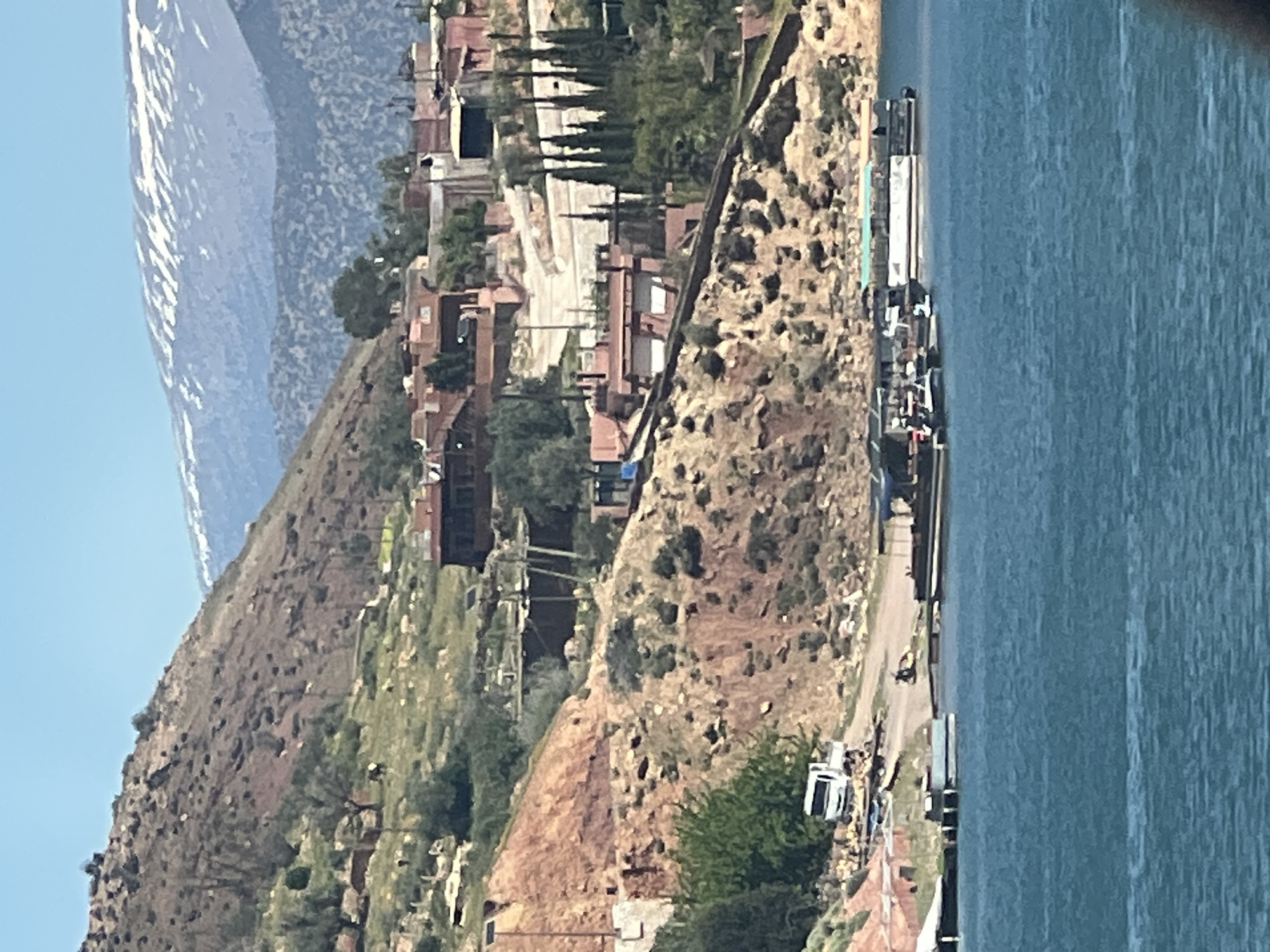The image presents a view of Bin El Ouidane, Morocco, likely taken from an elevated position. In the background, snow-capped mountains are visible under a clear sky. A hillside dominates the left side of the frame, gradually descending towards the city. The city itself is characterized by closely-packed terracotta-roofed buildings. A roadside path sits just off to the right, with parked vehicles visible along the road, as well as a body of water adjacent to the roadside.