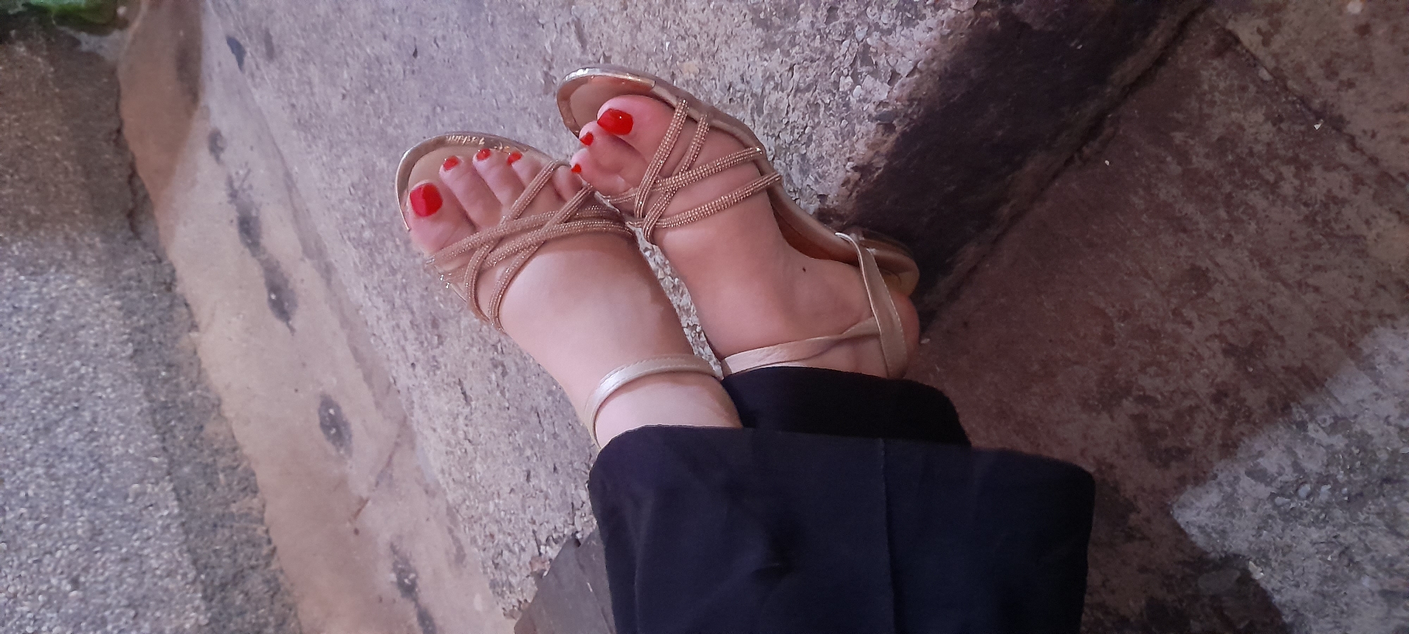 The image presents a close-up view of a person's feet adorned with gold-colored, strappy flat sandals. The toes feature bright red nail polish. The feet appear to be resting or placed on a weathered, textured concrete or stone surface, possibly steps or a low wall. Part of a pair of black trousers or pants is visible around the lower legs and ankles.
The setting appears to be outdoors, with the rough ground showing various shades of gray and brown, and some darker, possibly damp, spots. The lighting suggests it is daytime, with a soft illumination on the skin and sandals. No specific activity beyond the resting position of the feet is evident. There is no visible text in the image. The location is Juan Griego, Venezuela.