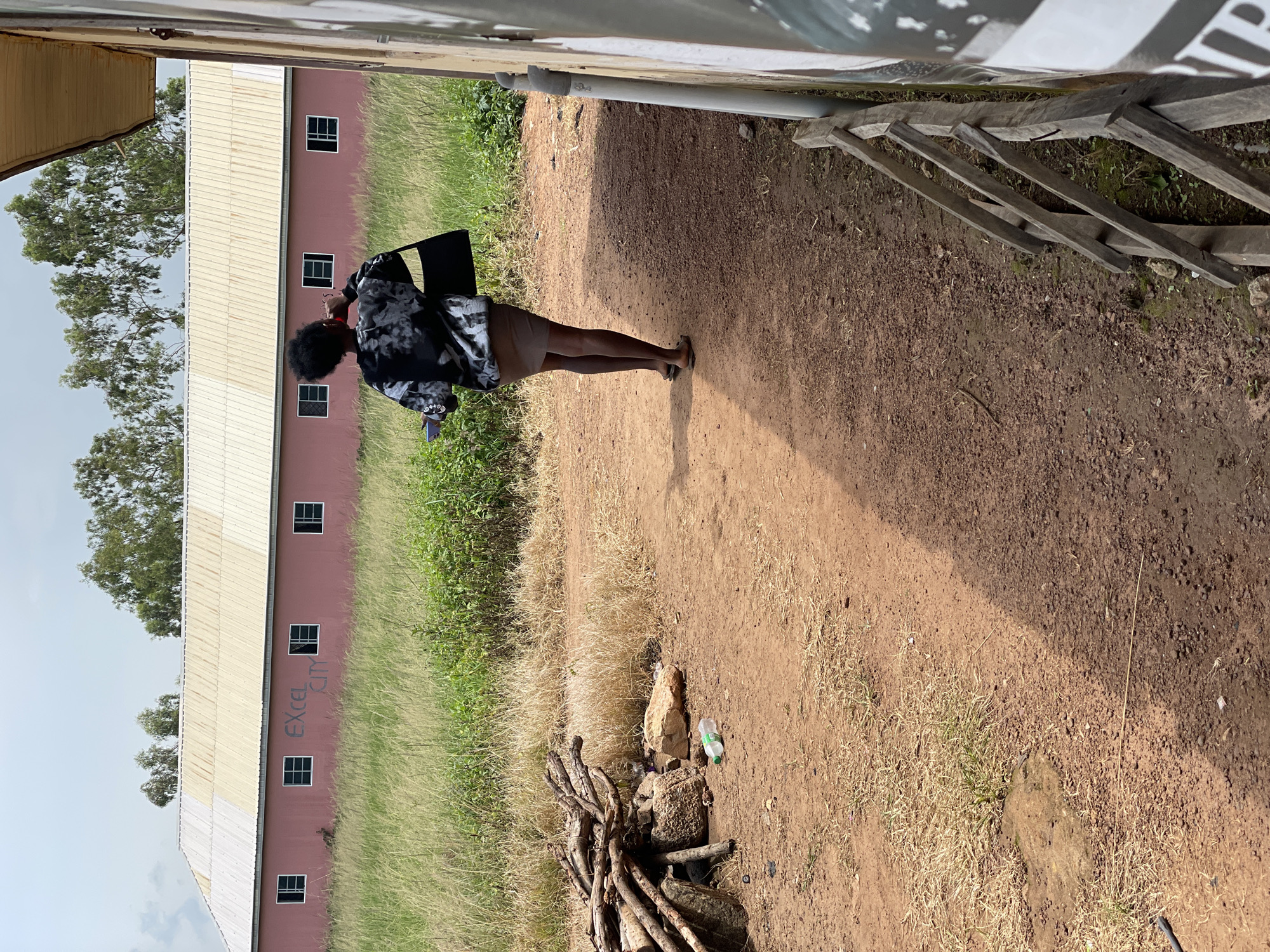 A woman walks away from the camera on a dirt path. She has dark curly hair and is wearing a black and white patterned long-sleeved shirt, a light brown skirt, and sandals. She is carrying a black purse on her right shoulder and holding a phone in her left hand. The path is lined with dry grass and scattered debris, including a pile of firewood and a plastic bottle. In the background, a long, pink building with several windows is visible, with the words "EXCEL CITY" spray-painted in grey on its facade. Trees and a cloudy sky form the backdrop. The scene appears to be in a rural or semi-urban area.