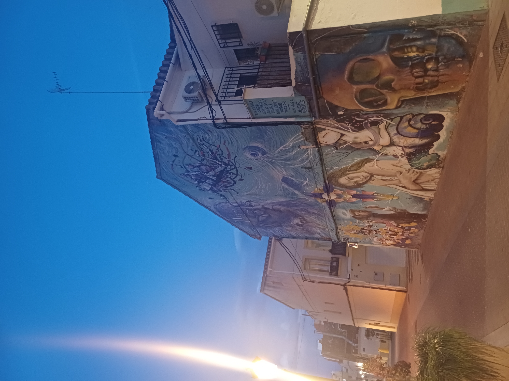 Here is a detailed description of the image:
**Scene:** A street scene in València, Spain, at dusk or dawn, with a wall adorned with a mural.
**Murals:** The mural, which is a primary focus of the image, is made of an interesting variety of images. It shows a skull, as well as a woman, and the name "José Segrelles" can be seen on the side of the wall.
**Buildings:** The architecture consists of white buildings with multi-story facades. Some buildings have balconies and air conditioning units.
**Environment:** The sky is a gradient of blue and orange, indicative of a sunrise or sunset. Street lighting creates a bright glare. Vegetation is visible on the side of the street.
**Objects:** Streetlights, antennas, and utility wires are also part of the scene.
**Overall Impression:** The image captures a blend of urban environment and artistic expression.
