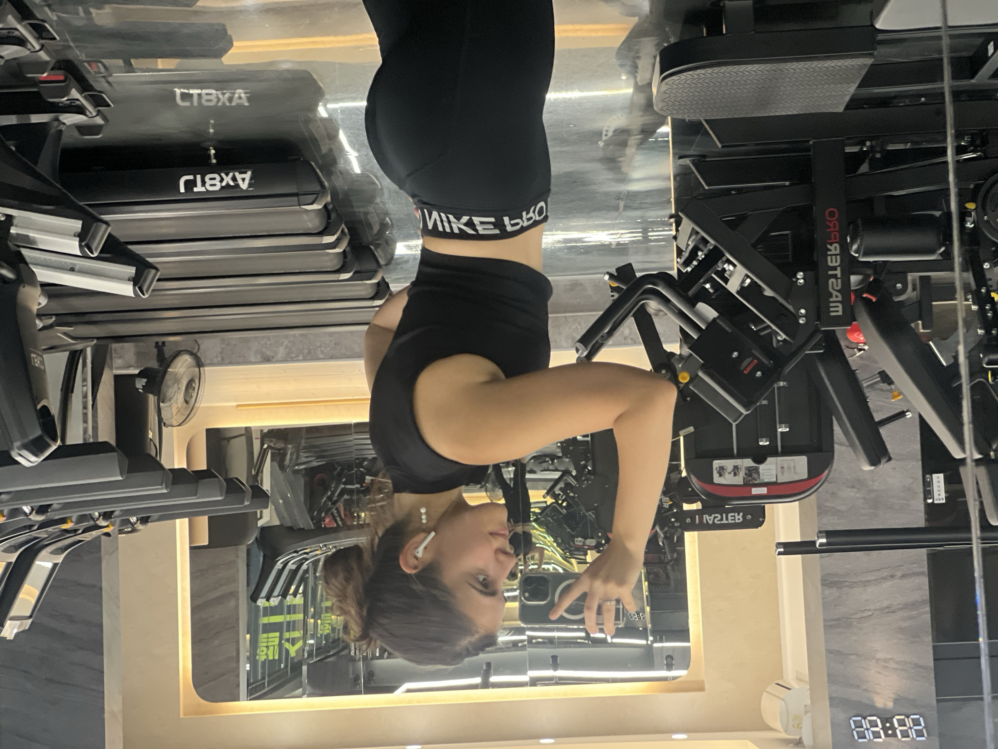 A young woman with dark hair pulled back into a bun stands in a gym and takes a selfie in a mirror. She is wearing a black sleeveless crop top and black leggings with a white logo band around the waist. She has white earbuds in her ears. Behind her, treadmills are lined up, and gym equipment is visible. A digital clock on the wall displays "00:00". The floor is reflective, showing the surrounding gym equipment.