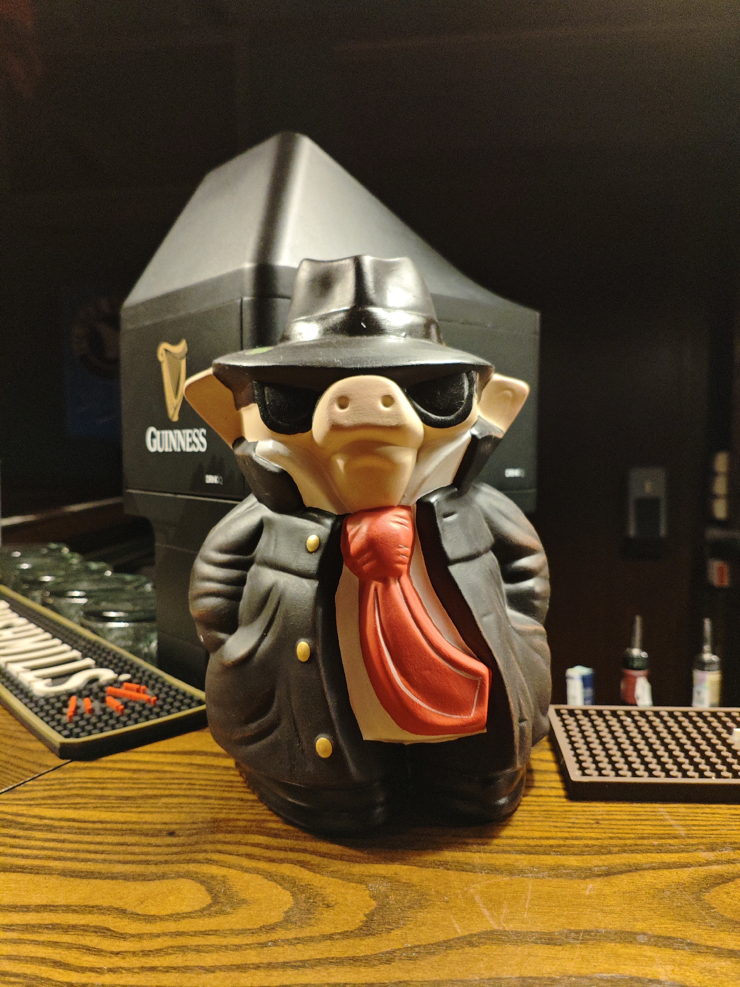 A ceramic pig figurine dressed as a mobster stands on a wooden bar top. The pig wears a black fedora, dark sunglasses, a black trench coat with gold buttons, and a bright red tie. Behind the pig is a black Guinness beer dispenser with the Guinness harp logo and text visible. To the left of the figurine is a black bar mat with white lettering that appears to say "GUINNESS" and some red plastic stirrers. To the right of the figurine is another brown bar mat with a grid pattern, and behind it, blurred bottles of liquor are visible. The setting is dimly lit, suggesting an indoor environment like a bar or pub. The overall mood is playful and somewhat mysterious, due to the figurine's attire. No specific time of day or weather conditions are discernible. The location context of Kyiv, Ukraine, is noted, but no direct visual cues in the image confirm this.