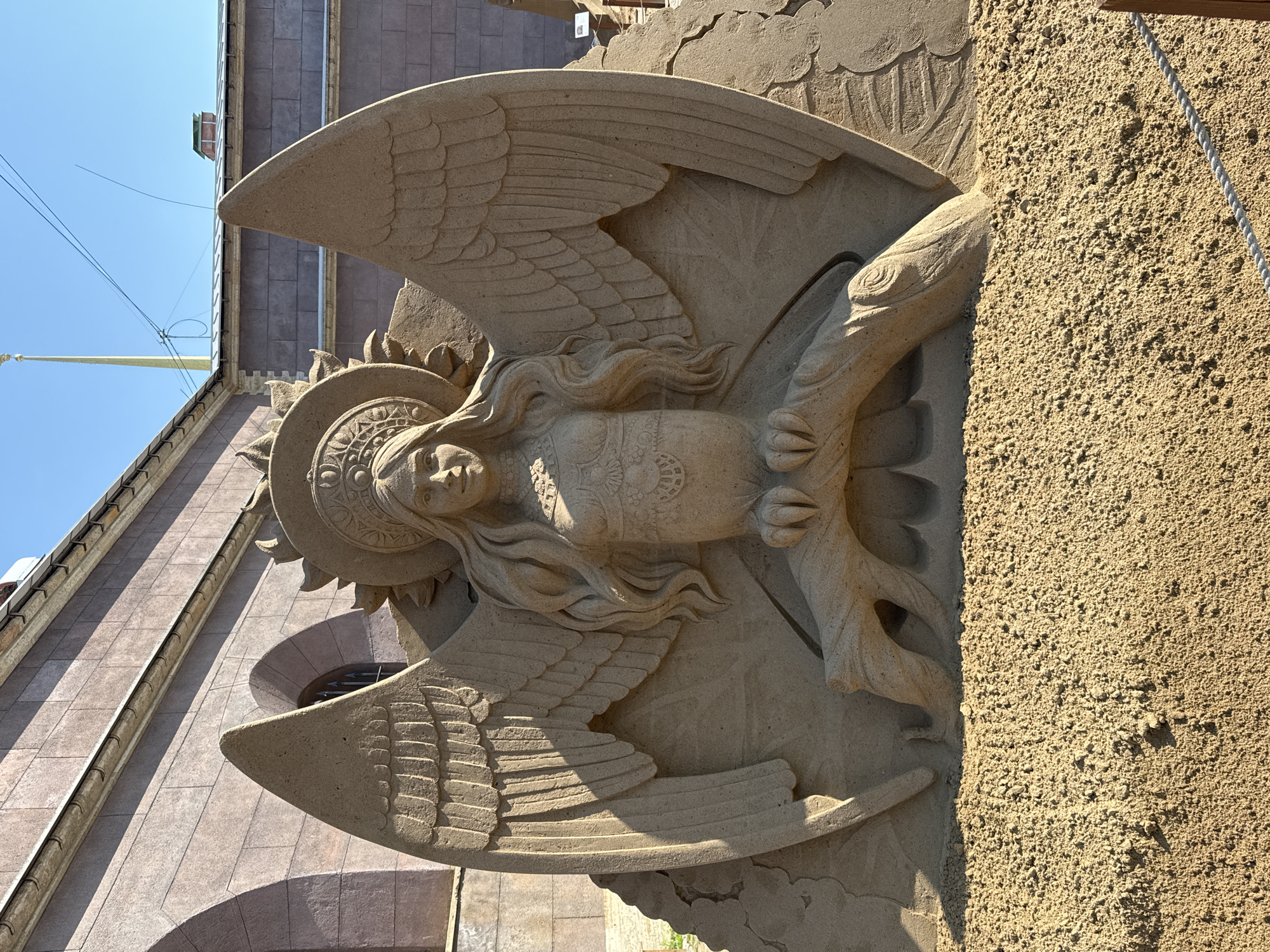The image shows a sand sculpture of a winged figure, possibly an angel or deity, with intricate details in the wings, hair, and face. The sculpture includes a sun-like halo behind the figure's head. The sculpture is located next to a building with a textured exterior. Judging from the style of the building and the sand sculpture festivals held there, it is likely in Saint Petersburg, Russia.