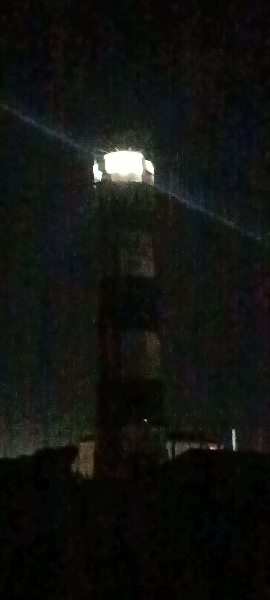 The image depicts a nighttime scene featuring a lighthouse. The lighthouse has a distinctive black and white striped pattern along its tower. A bright light emanates from the top of the lighthouse, illuminating the surrounding darkness. The lighthouse appears to be situated on a dark, possibly rocky or vegetated, terrain. The background is predominantly black, indicating it is night. There is a faint beam of light extending from the top of the lighthouse to the left, suggesting the direction of its illumination. The overall image quality is low, with visible noise and blur.