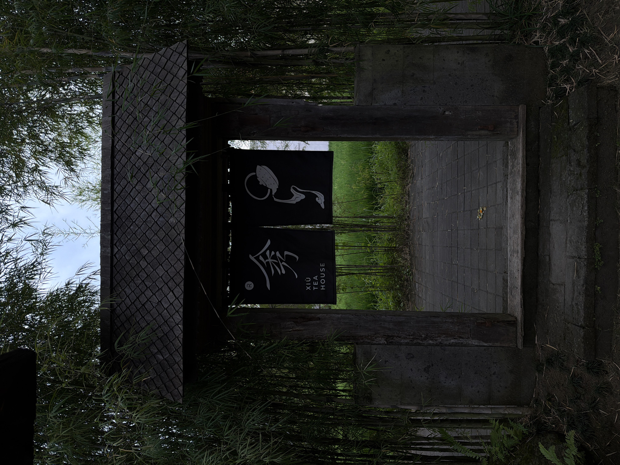 The image displays the entrance to Xiù Tea House, located in Singakerta, Indonesia. The central feature is a dark, gabled wooden gate structure with a prominent roof constructed from small, diamond-patterned dark shingles. This roof is supported by two robust, dark timber uprights, which are anchored by dark stone bases. Hanging from the overhead lintel are two black fabric banners. The left banner features a white stylized character above the text "XIÙ TEA HOUSE" in white capital letters, accompanied by a registered trademark symbol (®) in the upper left corner. The right banner displays a white illustration of a teapot pouring liquid.
The gate's threshold consists of a dark wooden beam, beneath which are several dark stone steps leading to a dark grey rectangular paver pathway. The entire structure is flanked by dense groves of tall green bamboo stalks and foliage, with some smaller green plants visible at ground level. Beyond the gate, an expanse of vibrant green vegetation, consistent with a cultivated field or paddy, is visible under a pale sky. A single yellow-orange deciduous leaf rests on the paver pathway immediately past the wooden threshold. No human figures are present within the scene. The lighting is diffused, suggesting an overcast day or heavily shaded environment.