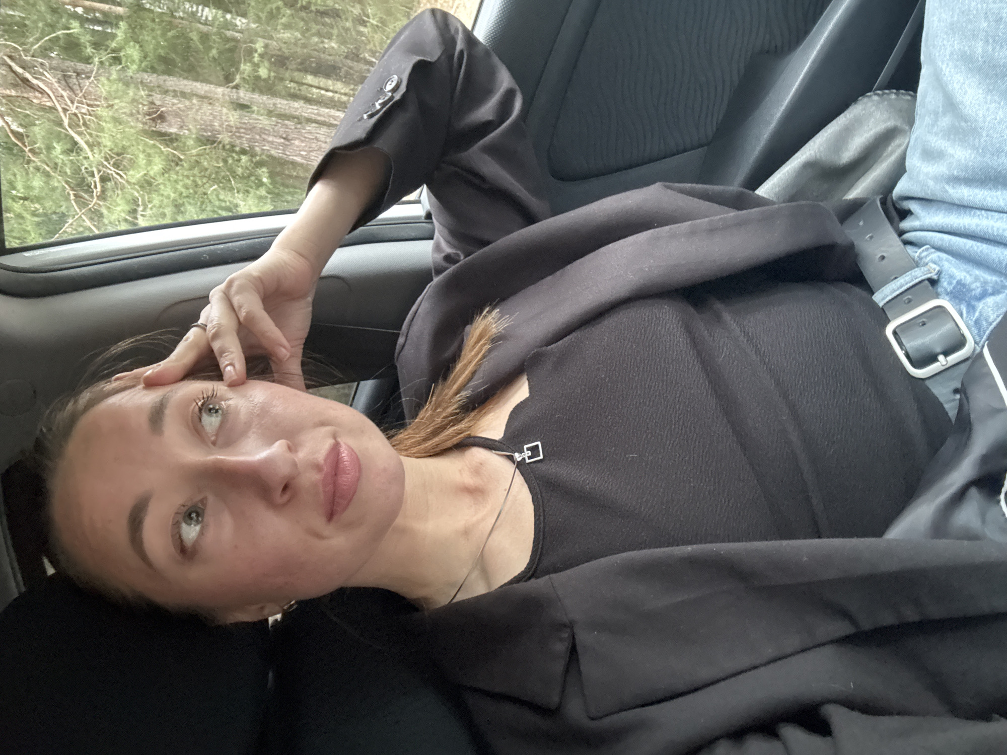 A young woman is captured in a close-up, low-angle shot, reclining or leaning back inside a vehicle, likely a car or van, in Chaikovsky, Russia. She is positioned with her head tilted to the left, her gaze directed upwards and slightly to her right, conveying a thoughtful or somewhat pensive expression. Her left hand rests gently on her forehead and hair, revealing a ring on her finger.
She is dressed in a dark, possibly black, short-sleeved top or dress, layered with a black blazer or jacket that is partially draped over her. A slender silver necklace adorned with a square pendant hangs around her neck. To the right side of the frame, a section of a blue denim garment, possibly jeans, and a black belt with a silver buckle are visible, suggesting the presence of another person or part of her own attire within the vehicle.
Beyond the vehicle's window, a natural outdoor environment is discernible, featuring a dense forest or wooded area with visible trees and foliage, blurred by movement or focus. The ambient light indicates a daytime setting. Elements of the car's interior, such as a dark fabric seat and the window frame, are visible. The image conveys a moment of contemplation or rest during travel. There is no visible text in the image.