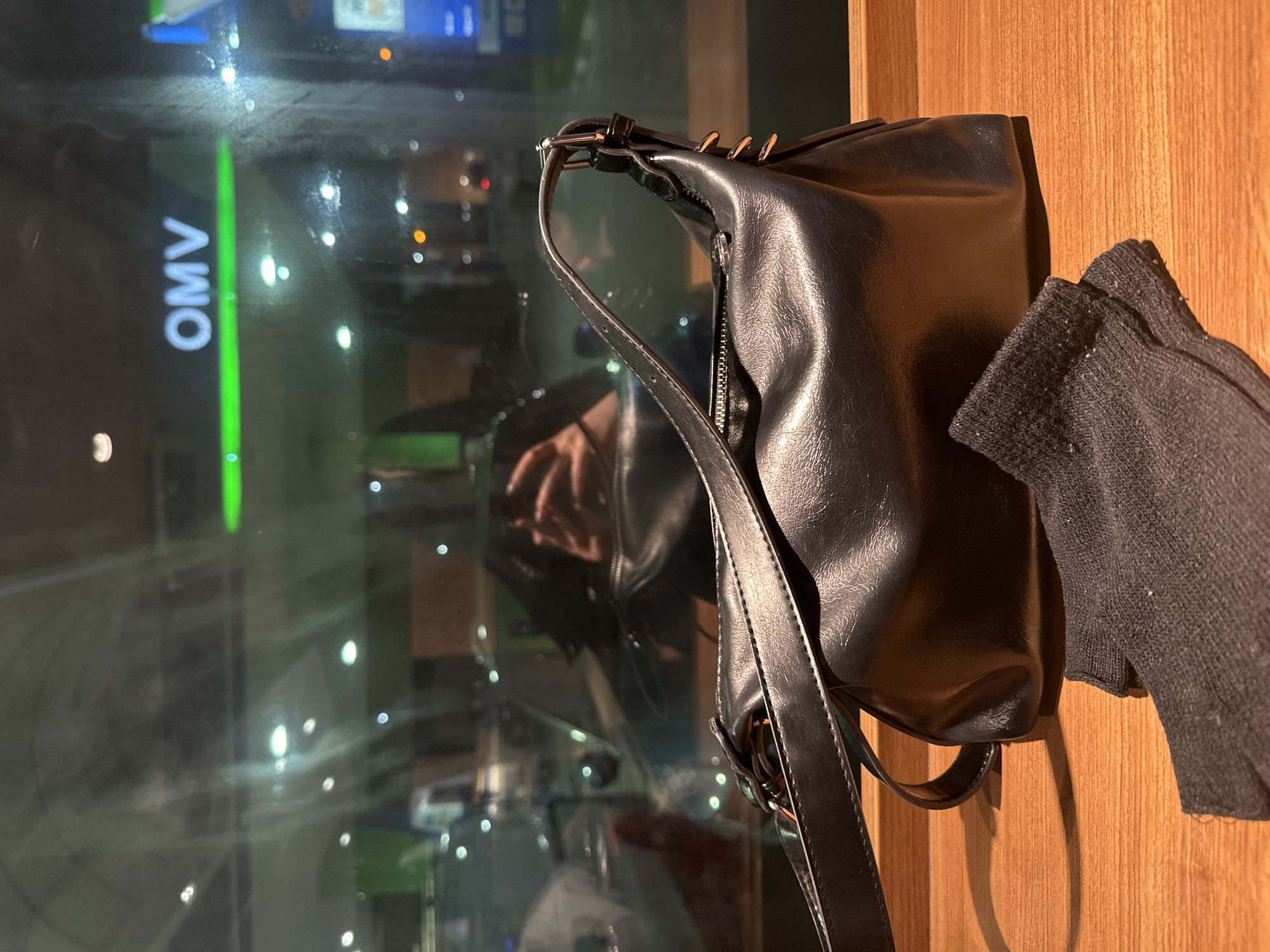 A black handbag with a shoulder strap sits on a wooden surface next to a black sweater or article of clothing. Reflected in the window behind the bag are the lights and signs of Stara Zagora, Bulgaria, including a clear reflection of an "OMV" gas station sign. The time of day appears to be evening or night.