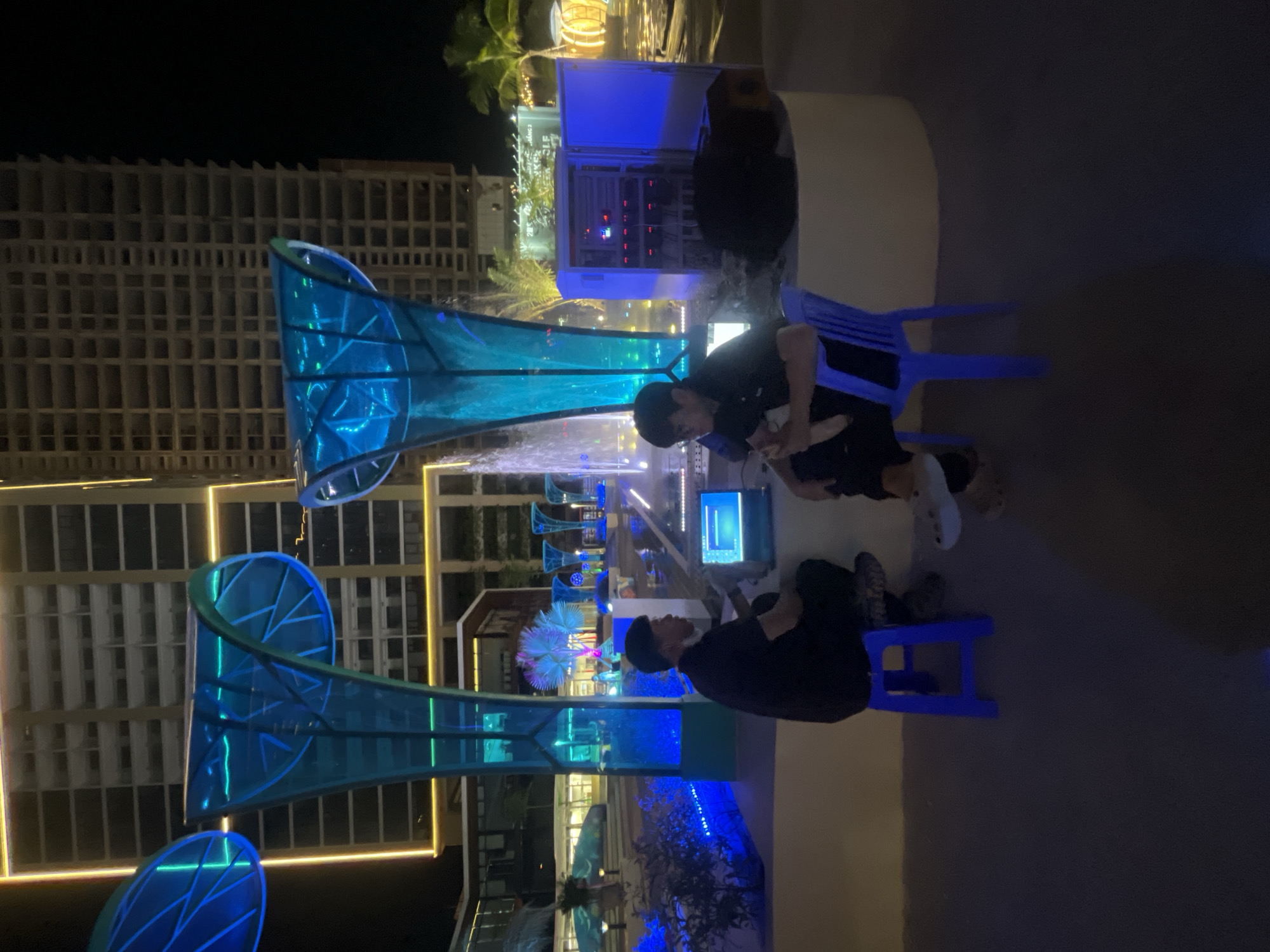 The image depicts an outdoor scene at night, likely in Đà Nẵng, Vietnam, featuring an urban setting with modern architectural elements. In the foreground, two individuals are seated on blue stools, facing each other and appearing to interact with a laptop placed on a low surface between them. One person is wearing a black t-shirt and a black cap, while the other is in a dark polo shirt and glasses.
Dominating the background are large, illuminated structures with a leaf-like design, glowing with vibrant blue and green light. These structures appear to be part of an artistic installation or a decorative element for a public space. Behind them, a tall, unfinished building with multiple floors and visible concrete framework suggests ongoing construction or development. Fountains are also visible, adding to the ambiance of the location, and palm trees can be seen to the right, indicating a tropical or subtropical climate.
The scene is illuminated by a combination of the colorful artistic lights, the building's exterior lighting, and ambient artificial light sources. The ground surface is paved, and the overall impression is that of a modern, possibly recently developed, urban public space. A control panel with multiple indicators is positioned to the right of the individuals, suggesting some form of technical operation or management of the surrounding installations. The presence of the laptop and control panel implies that the individuals might be involved in setting up, testing, or operating these illuminated features or fountains.