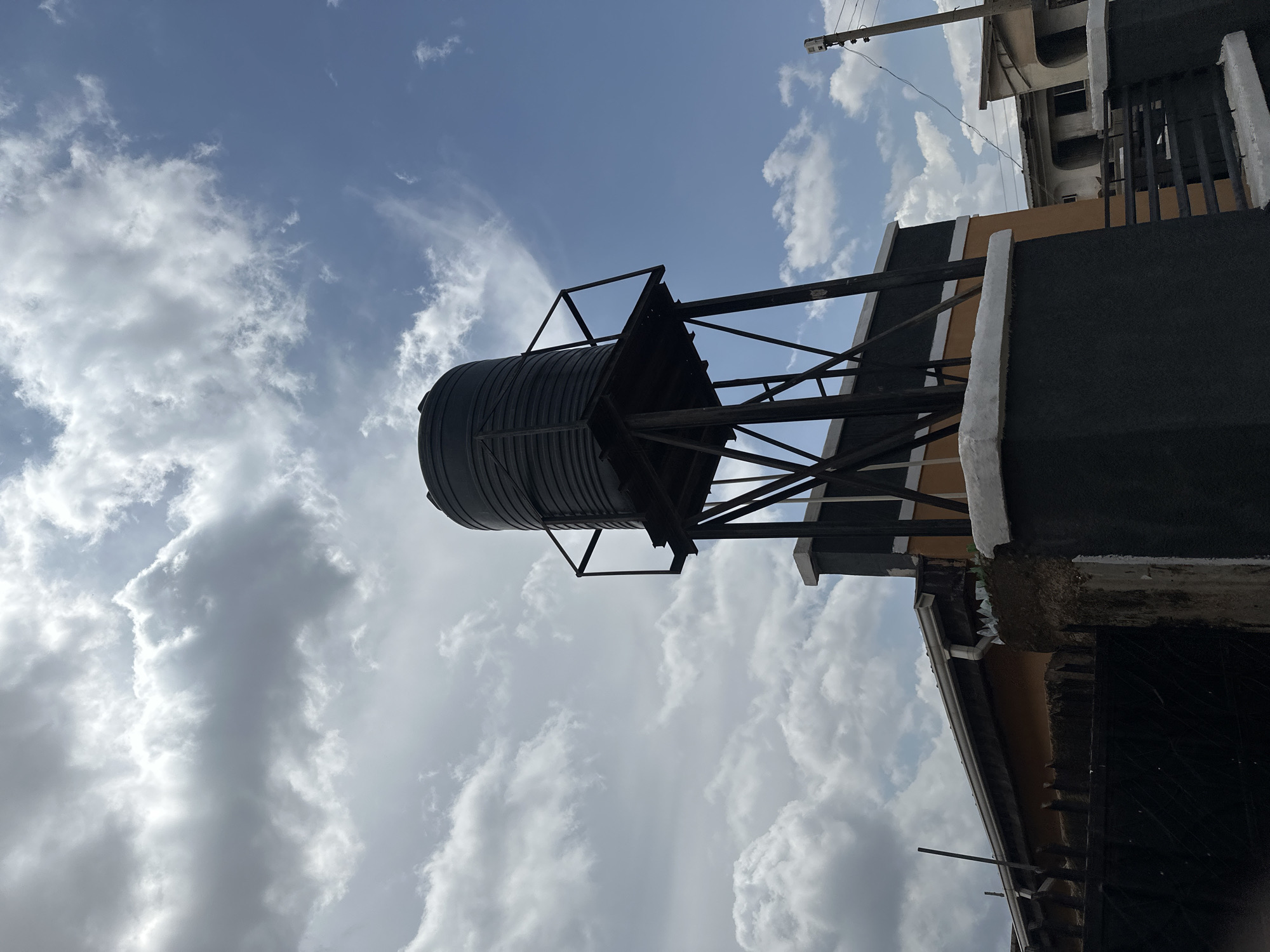 A low-angle, medium shot captures a black water tank elevated on a metal platform, against a backdrop of a partly cloudy sky with blue patches. The tank is situated above a building with an orange and grey facade. In the foreground, a dark metal fence and a grey concrete pillar are visible. The scene appears to be outdoors in a residential area, possibly in a city, though no specific location details are discernible in the image.