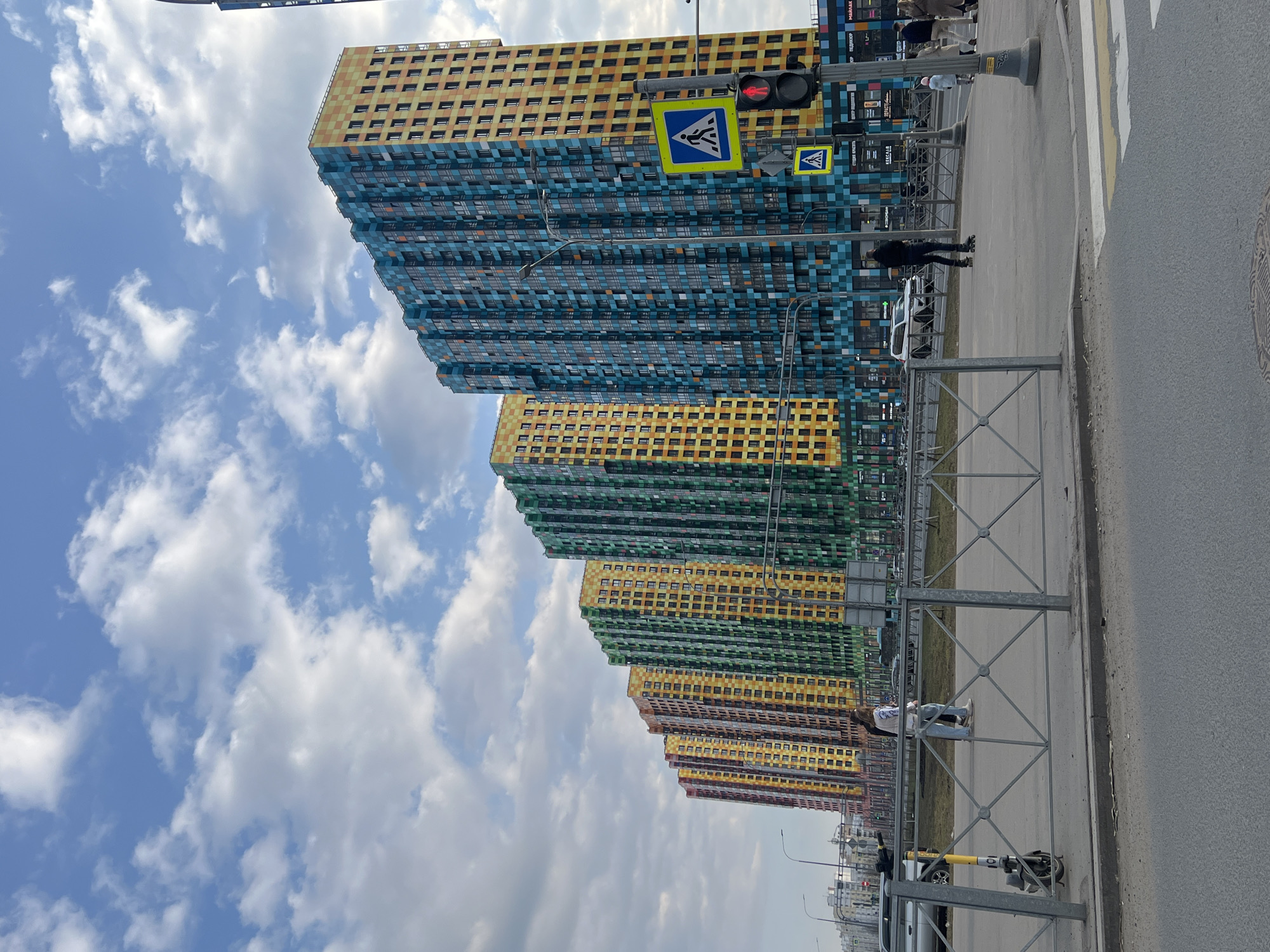 The image depicts a row of colorful, high-rise apartment buildings in the Kudrovo district of Saint Petersburg, Russia. The buildings display a variety of colors, including blue, yellow, green, and orange. The sky is blue with scattered white clouds. A street with a pedestrian crossing, traffic lights, and a metal fence runs parallel to the buildings. People can be seen walking along the sidewalk. Several scooters are parked near the fence. A paved road is visible on the right side of the frame.