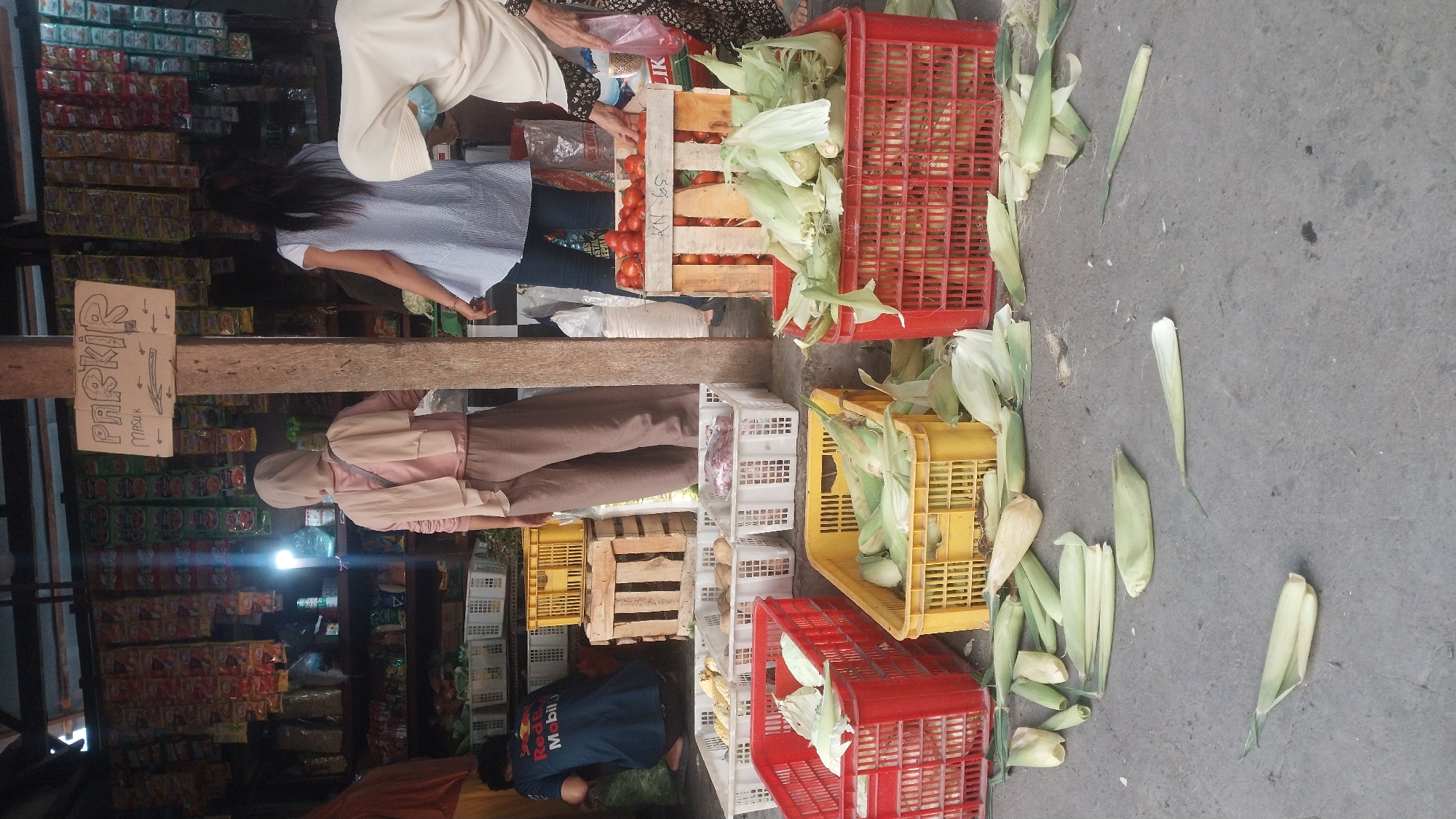 A street market scene unfolds in Keboananom, Indonesia, featuring a variety of fresh produce and shoppers. In the foreground, several red and yellow plastic crates are filled with corn, some with husks still attached, while discarded husks and corn cobs are scattered across the concrete ground. Behind the produce, white plastic crates hold more vegetables, and a wooden crate contains red tomatoes.
Three individuals are visible, two women and one man, interacting within the market. A woman in a light-colored headscarf and a pink tunic is partially obscured by a wooden post, while another woman in a blue and white striped shirt and jeans stands facing away from the camera, towards a stall. A person wearing a dark blue shirt with red and white lettering is crouched down behind a display of produce on the left. Overhead, a sign with "PARKIR" and arrows is attached to a wooden support. The background reveals shelves stocked with various packaged goods, suggesting a small, bustling market environment. The lighting suggests it is daytime, likely overcast or shaded by awnings.