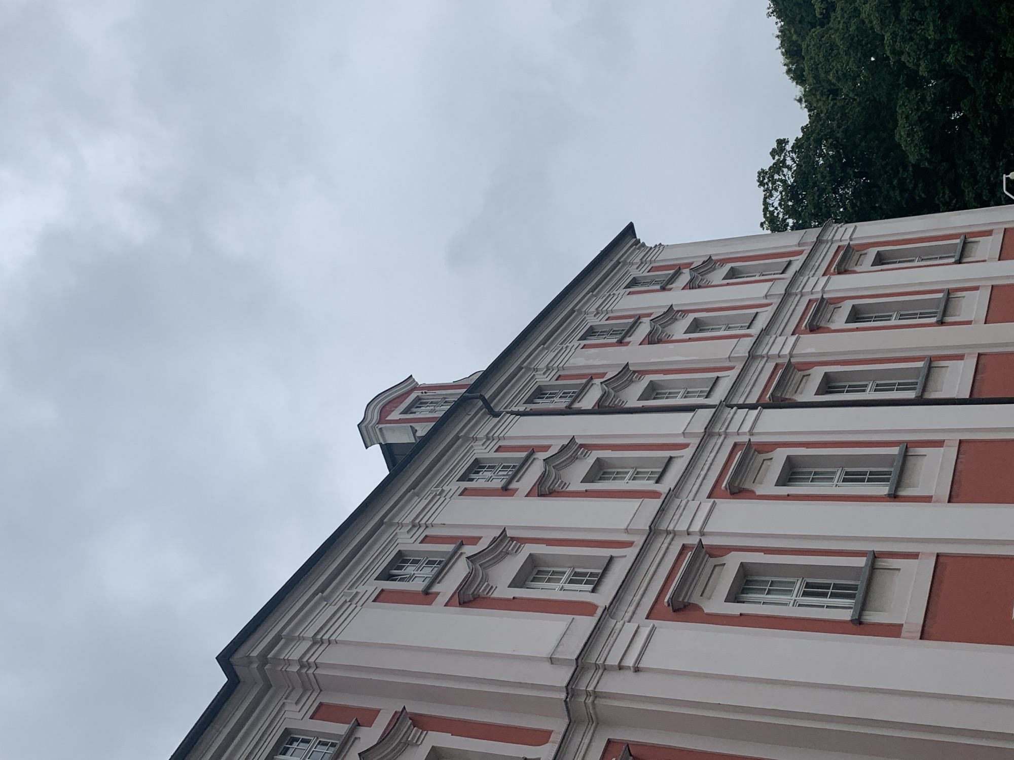 The image shows a low-angle exterior shot of the side of a pink and white building with a dark grey, cloudy sky. The building is ornate with white trim around red sections and decorative window frames. It has multiple stories with many windows. Green trees are visible on the right side of the frame, obscuring a portion of the sky and the building's facade. The building is likely the Imperial Castle (Zamek Cesarski) in Poznan, Poland, given its architectural style.