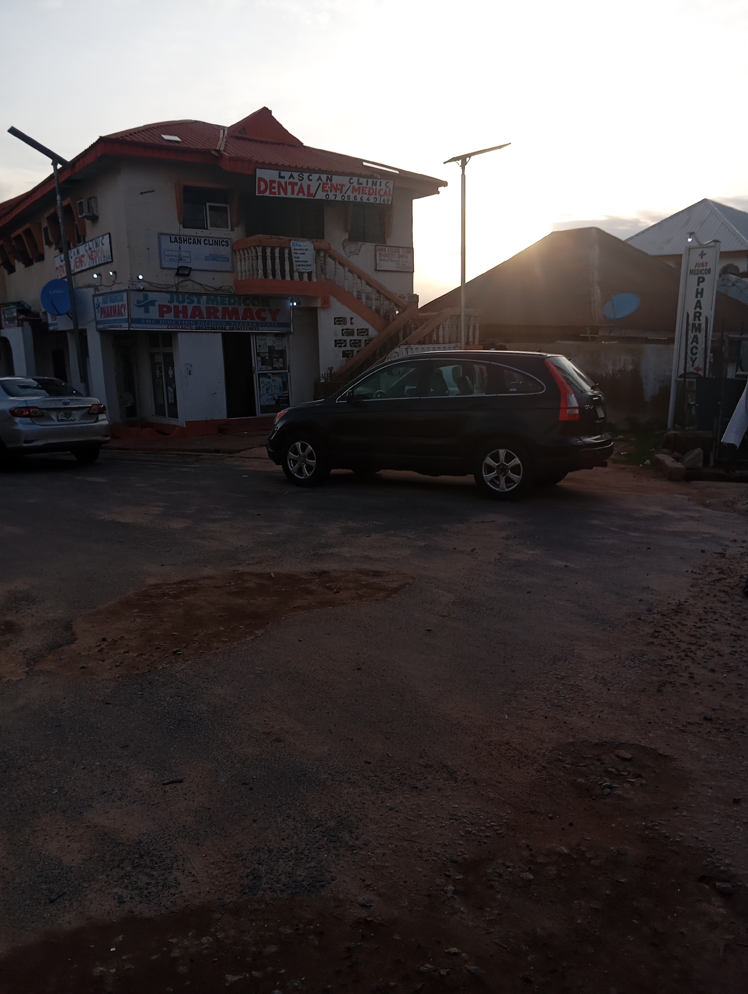 The image shows a street scene in Jalingo, Nigeria, with several buildings and vehicles. In the foreground, a black SUV is parked on the right side of the road, and a silver sedan is partially visible on the left. The buildings appear to be commercial establishments, with signs indicating "LASHCAN CLINICS," "DENTAL/ENT/MEDICAL," and "JUST MEDICOR PHARMACY." One sign for "JUST MEDICOR PHARMACY" is a vertical banner on the right side of the image. The sky is overcast, suggesting it might be early morning or late afternoon. The road surface is asphalt with some patches of dirt and puddles. The overall impression is of a typical urban street in Nigeria.
