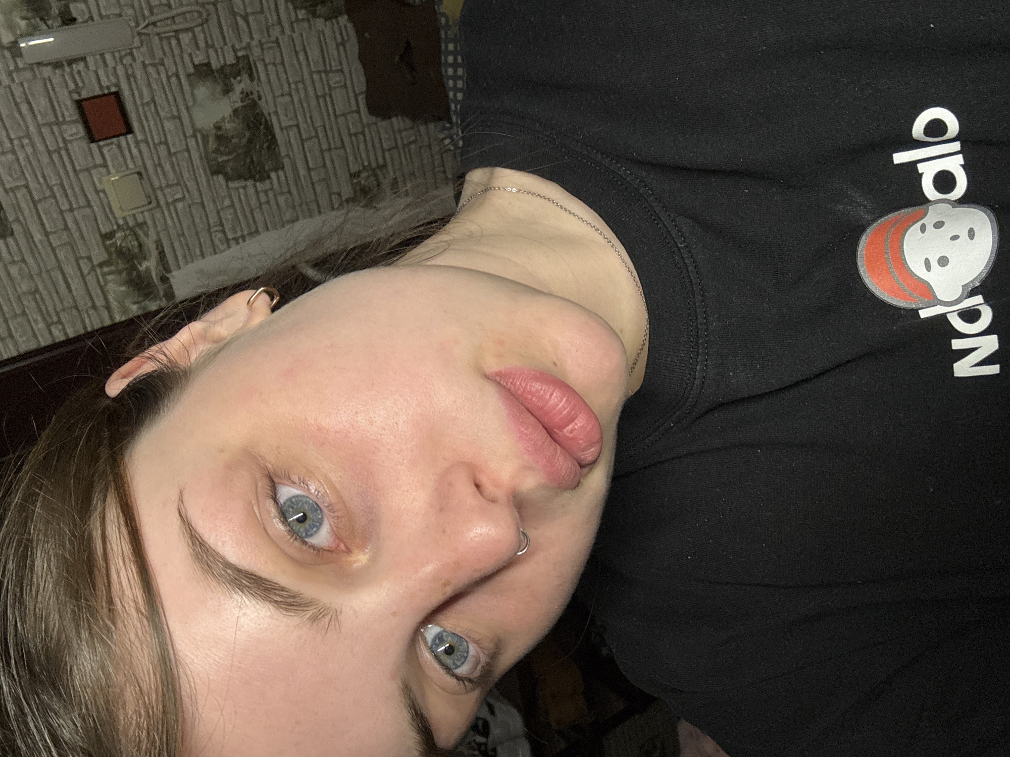 A close-up portrait shows a young woman indoors. The woman has light skin, blue eyes, and light brown hair. She has a piercing in her nose. She is wearing a black sweatshirt with a cartoon character printed on it. A thin silver chain is visible around her neck. The background includes a textured wall, with a wooden frame and a red item on it. No specific information indicates this picture was taken in the Новосельское сельское поселение, Ukraine.