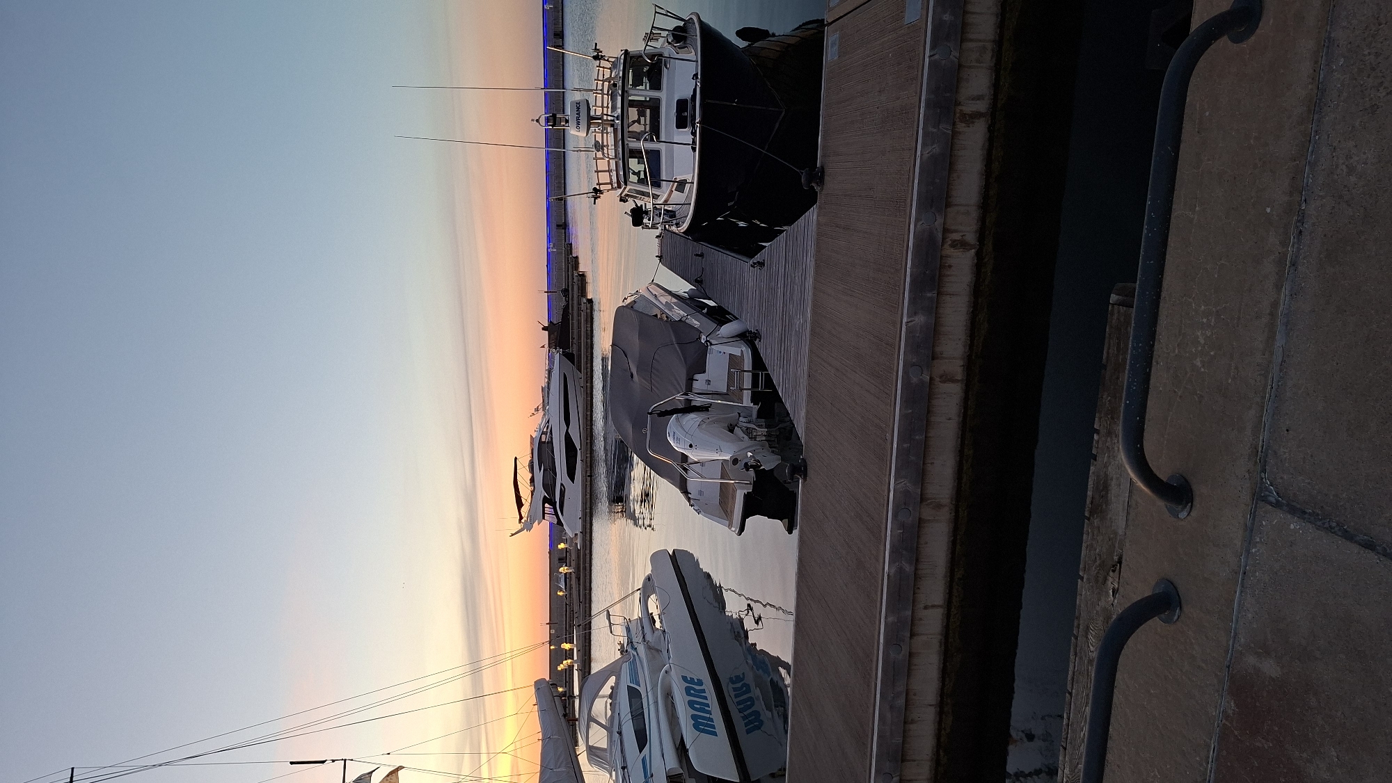 The image depicts a marina in Tallinn, Estonia, during a calm sunset. Several boats are docked at wooden piers, with a clear sky transitioning from pale blue to soft orange and pink hues. In the foreground, a concrete pier with dark metal railings lines the bottom of the frame. Beyond it, a wooden dock hosts several vessels. To the left, a white catamaran-style boat with "MARE" written in blue lettering is moored. Next to it, a smaller boat is partially covered with a dark gray tarp, revealing a white outboard motor. To the right, a dark-hulled boat with a white superstructure, identified by the "LOWRANCE" logo on its radar dome, is docked. In the mid-ground, a sleek white yacht with a dark top is also visible. The water is still, reflecting the soft colors of the sky and the boats. The overall atmosphere is serene and peaceful, characteristic of a marina at dusk.