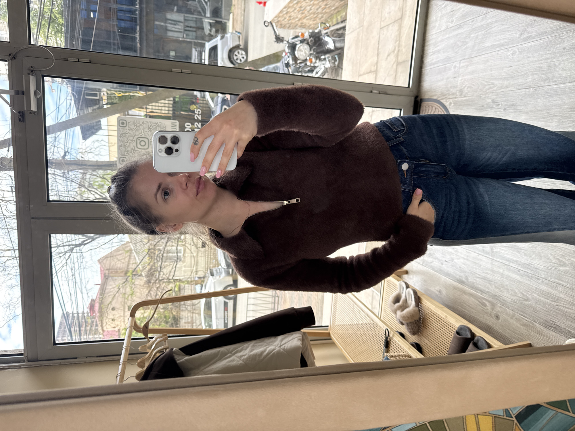 A young woman is captured taking a mirror selfie in an indoor setting, likely an entryway or dressing room, characterized by a light-colored wooden floor and large windows. She is wearing a brown, fuzzy, long-sleeved top with a quarter-zip neckline and blue denim jeans. Her dark hair is pulled back, and she has light-colored eyes and pink nail polish. She holds a white iPhone with multiple cameras in her left hand, facing the mirror, while her right hand is casually tucked into her jeans pocket.
The reflection in the mirror shows a view outside through the windows, revealing what appears to be a street scene. A silver car and a classic motorcycle are visible on the street, alongside buildings and trees, some with bare branches and others showing early signs of foliage, suggesting spring or late winter. The natural light indicates it is daytime.
To her side, within the room, there is a simple wooden clothes rack with hangers and garments, including a quilted white item. Below the windows, a light-colored wooden furniture piece with a woven rattan front holds several pairs of slippers – one fuzzy beige pair and one dark brown pair. A small item, possibly a keychain, hangs from this furniture. A partially visible patterned rug or mat is on the floor. On the glass, near her head in the reflection, a QR code sticker is visible, along with fragmented text that appears to include "00" and "12 25", and the letters "UMI". The general architecture and outdoor scene are consistent with an urban environment like Batumi, Georgia.