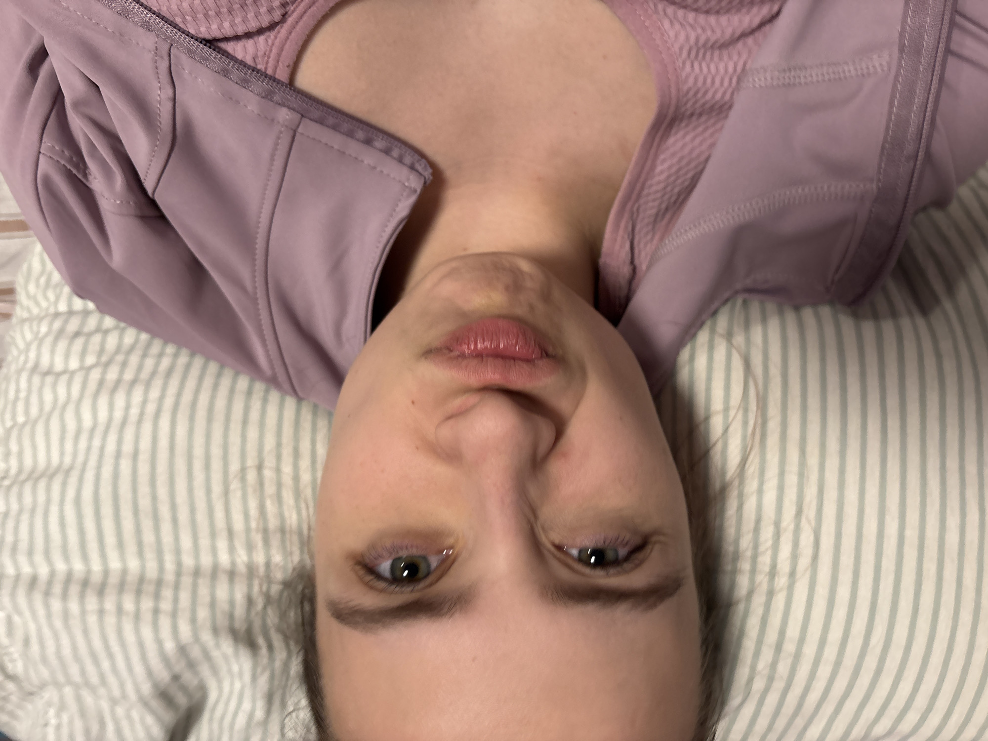 The image is a close-up, overhead portrait of a person, likely a young adult female, lying down. The photograph is oriented with the subject's head towards the bottom and her body extending upwards.
The subject has fair skin, light-colored eyes that appear greenish-hazel, and dark eyebrows. Her lips are slightly parted, revealing a natural pink color, and her expression is neutral and calm. Her dark hair is mostly out of frame, seen only at the edges of her head where it rests on a pillow.
She is dressed in a light purple or mauve athletic jacket or top. The garment features visible stitching details, particularly across the chest and shoulders, suggesting a structured or paneled design. The exterior fabric appears smooth, while a ribbed, lighter pink or mauve material is visible underneath the jacket, likely an inner layer or tank top.
The person is resting on a pillow that has a pattern of alternating light green and white vertical stripes. The lighting in the image is soft and diffused, indicating an indoor setting. No specific location cues are visible beyond the pillow and clothing, nor is there any visible text. The overall impression is one of a relaxed, casual moment.