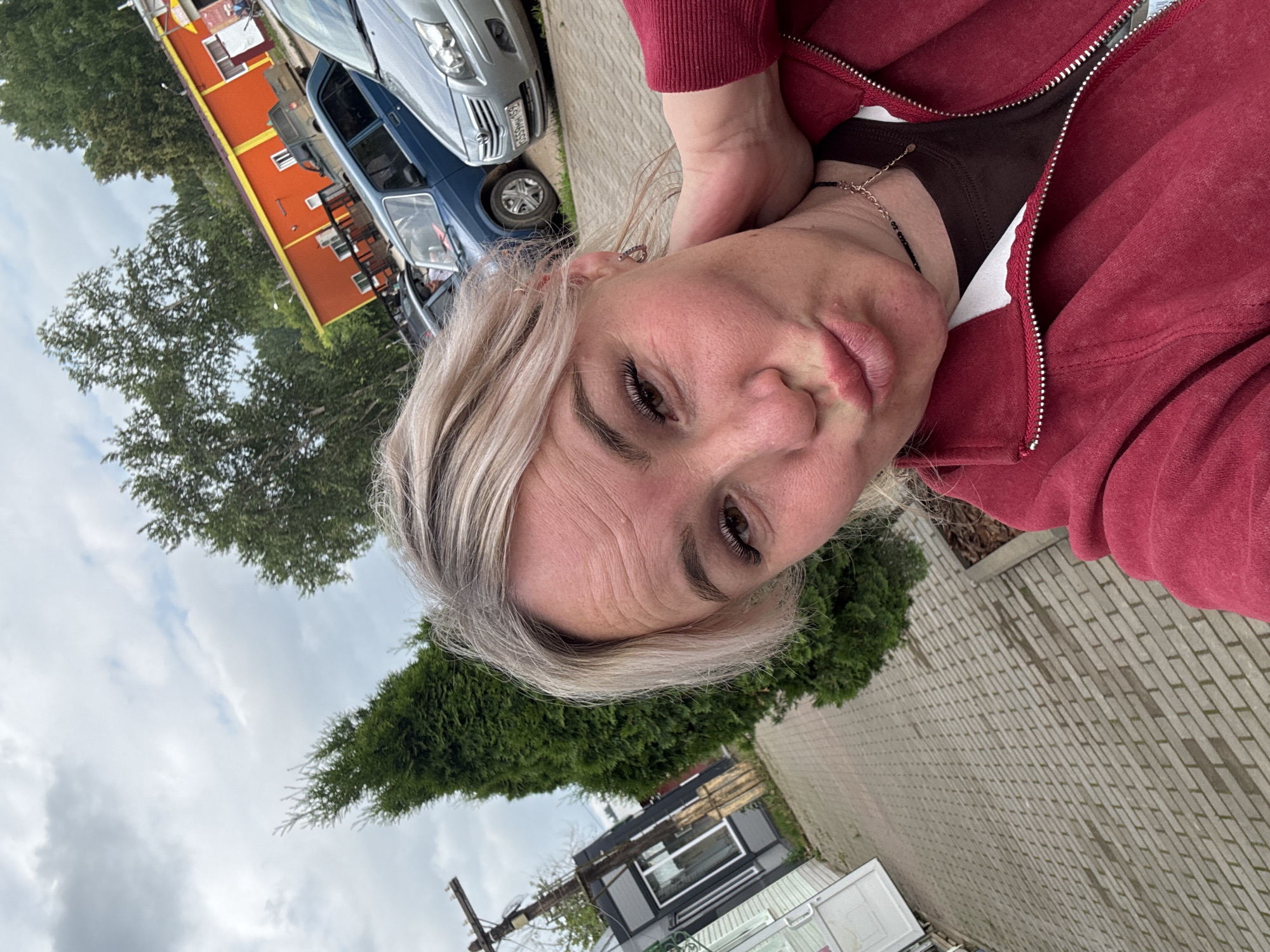 An outdoor, eye-level, medium shot captures a woman with blonde hair and light skin taking a selfie. She is wearing a dark brown t-shirt under a red zippered hoodie and a thin necklace. Her head is tilted, her right hand is touching her neck, and her lips are pursed. In the background, a tall, dense evergreen tree stands to her left, followed by a paved walkway. Beyond that, there is a gray building with a white door and windows, and a utility pole. Further in the distance, several cars are parked in front of an orange building with a yellow trim. The sky is overcast with clouds. The scene is likely in a public area, possibly a parking lot or a street in Chastyye, Russia.