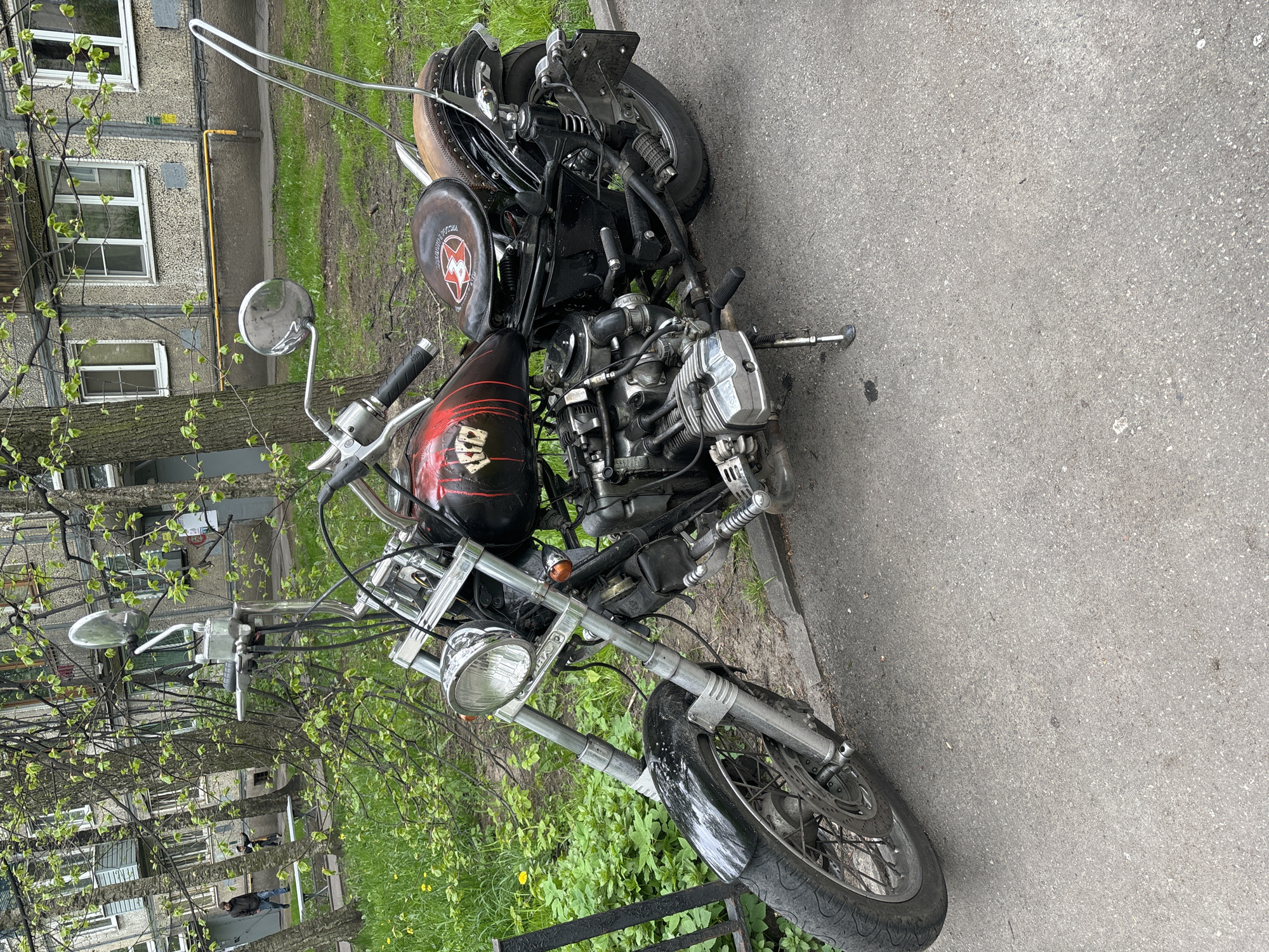 The image shows a custom motorcycle parked on an asphalt surface alongside a curb. The motorcycle appears to be a heavily customized chopper style bike, with a black and red gas tank featuring white skull graphics. It has a single seat with a brown leather trim and a custom sissy bar. The engine is exposed, appearing to be a boxer type engine. The motorcycle is parked on a sidewalk near a building, with green foliage and trees in the background. There are two people walking down the sidewalk in the background. Given the style of buildings and architecture in the background, the location is likely in Saint Petersburg, Russia.