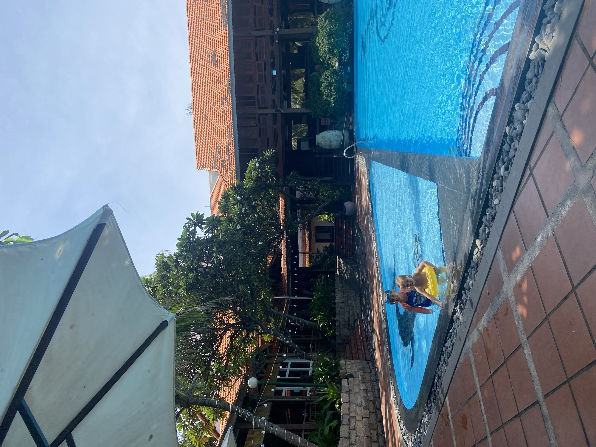 Two children are in a bright blue swimming pool near the edge. The child on the left, wearing goggles and a rash guard, is standing. The child on the right is sitting in the water with a yellow inflatable ring. In the background, there is a building with a red tiled roof, lush green trees, and string lights. The area around the pool is paved with terracotta tiles and decorative stones. A large white umbrella is visible in the upper left corner. The scene appears to be a resort or hotel in Phu Thuy Ward, Vietnam, given the architectural style and vegetation.
