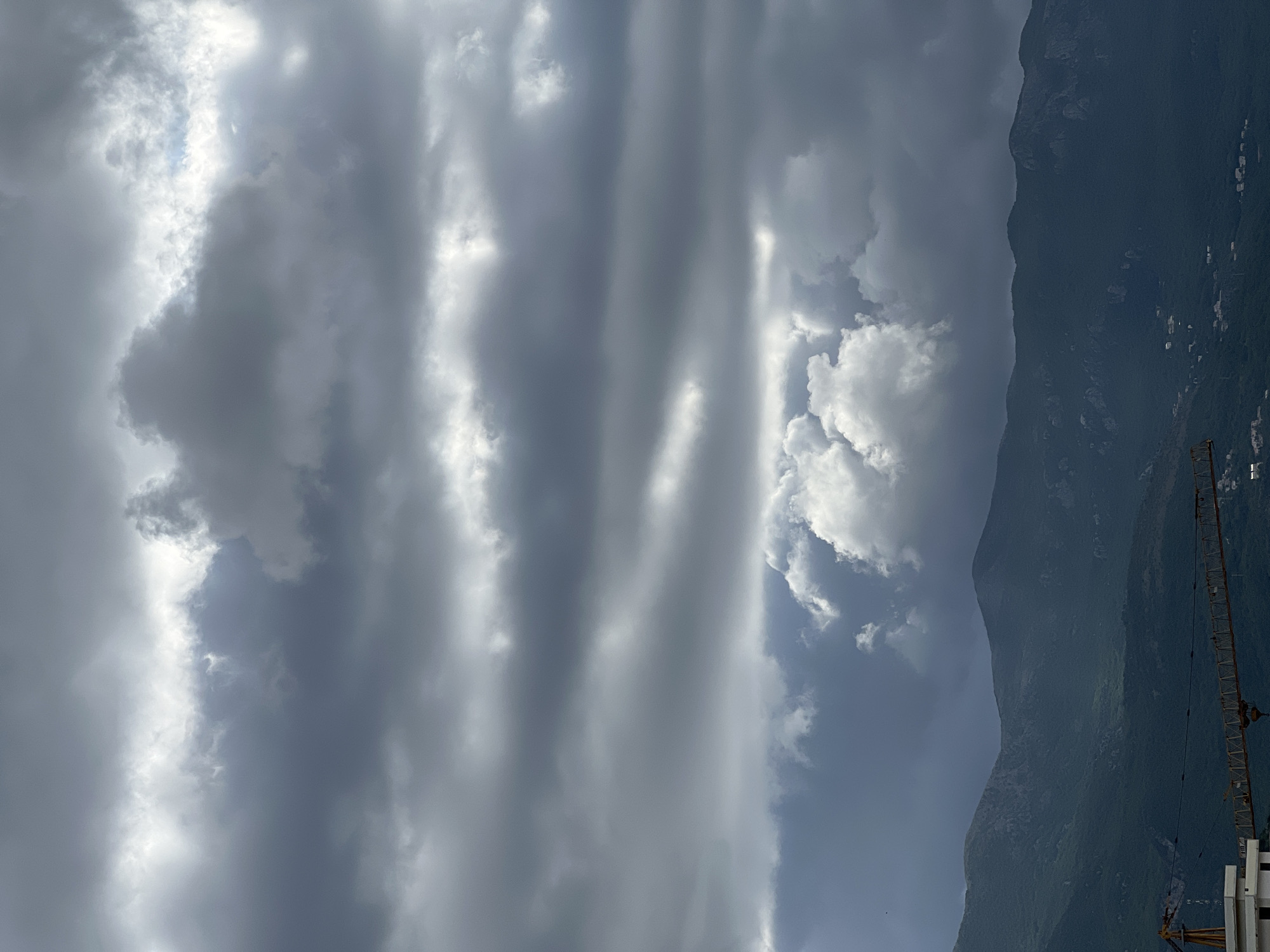 The image shows a view towards a tall mountain in what appears to be Budva, Montenegro, under a sky filled with dramatic, gray clouds. The clouds are layered and textured, with some areas allowing sunlight to break through. A crane is visible on the right side of the image. The mountain is dark and steep, with some visible structures or settlements.