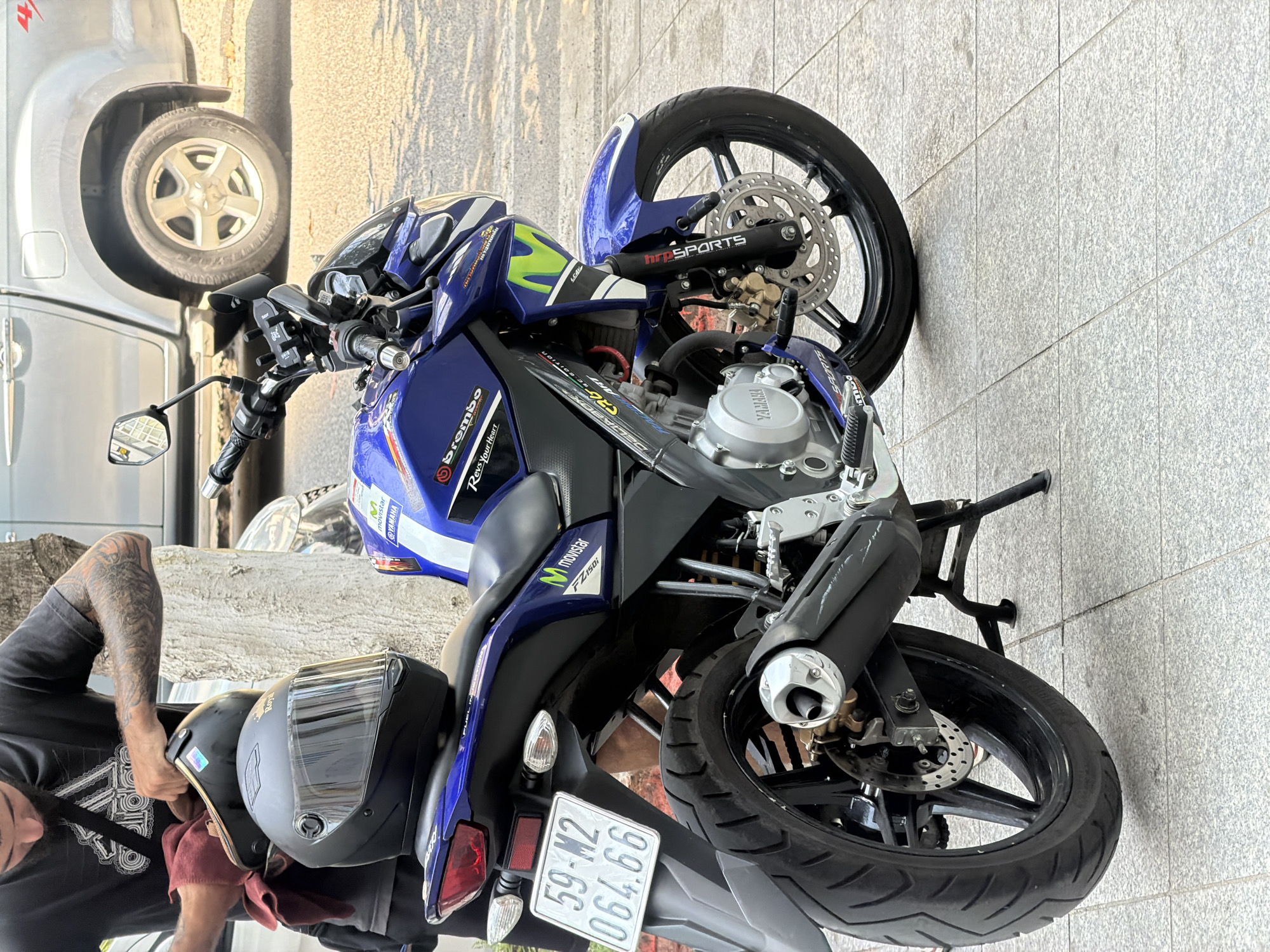 A man with tattooed arms is seen cleaning a motorcycle helmet that is resting on the seat of a blue and white Yamaha motorcycle. The motorcycle is parked on a tiled sidewalk in front of a silver pickup truck. The license plate of the motorcycle reads "59-M2 064.66". The scene appears to be in an urban setting, possibly Bac Nha Trang Ward, Vietnam, given the context. The overall lighting suggests it is daytime, with shadows indicating sunlight.