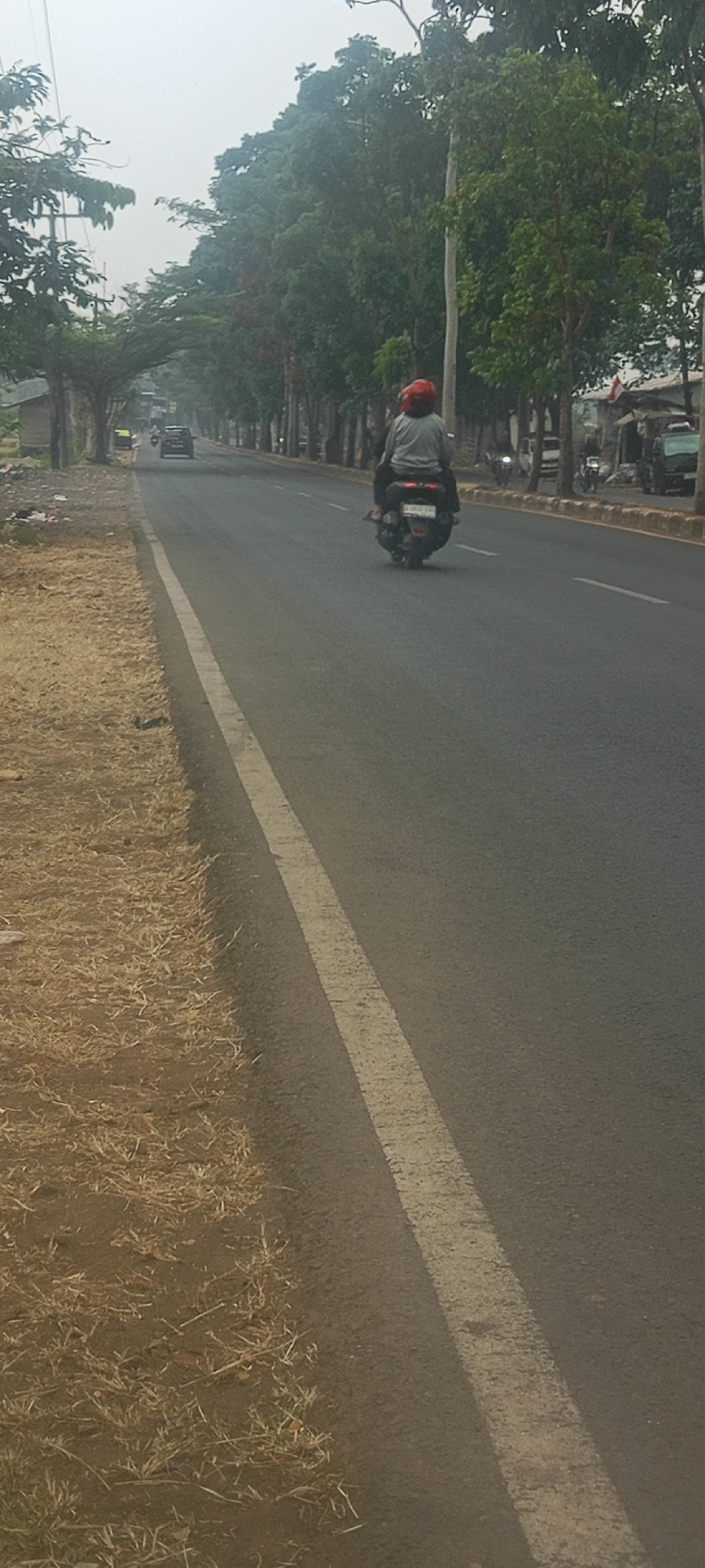 A motorcyclist wearing a red helmet and a gray long-sleeved shirt is riding a dark-colored scooter down a paved road in Soreang, Indonesia. The road has a white line marking the lane. The motorcyclist is facing away from the camera. Trees line the right side of the road, some appearing to form an archway over the road further up. The left side of the road has dry grass and dirt. Other vehicles are visible in the distance on the road. The sky is overcast, suggesting it may be a hazy or cloudy day.