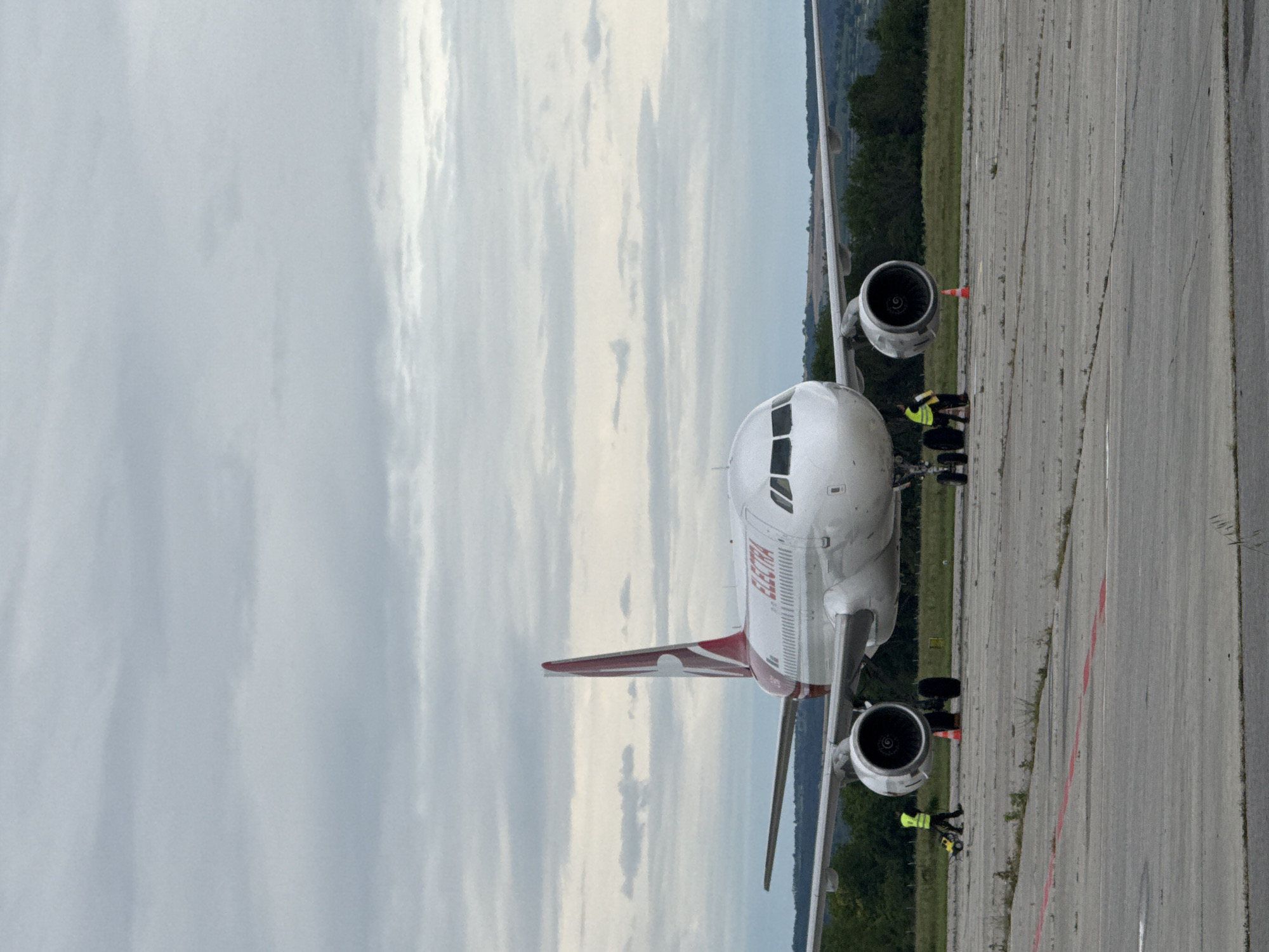 An airplane with "ELECTRA" written on its side is parked on an airport tarmac. The airplane is white with a red tail fin. Two individuals in yellow vests are visible near the front landing gear, one appearing to operate a yellow cart. Orange cones are placed on the tarmac near the wheels. The background consists of a cloudy sky and a treeline with rolling hills. The scene is located at an airport in Aksakovo, Bulgaria.