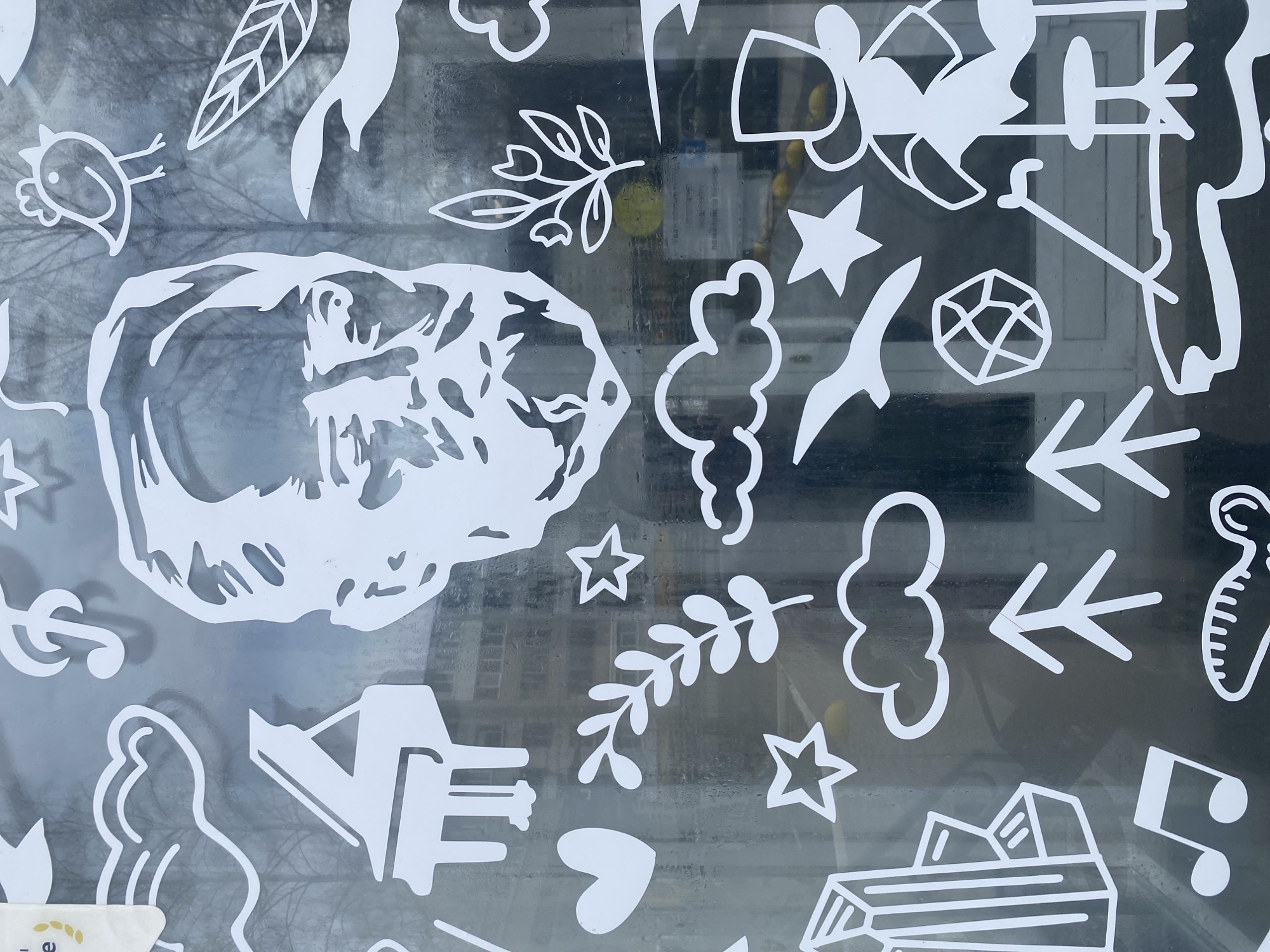 A view through a window covered in white paper cutouts, likely in Chaikovsky, Russia. The cutouts include a bird, leaf, plant sprigs, stars, clouds, arrows, a geometric shape resembling a geodesic dome, musical notes, a silhouette of a building, and an abstract organic shape. The background visible through the window is a blurry urban landscape with trees and buildings. The window is slightly fogged or has water droplets on it. The overall scene suggests a whimsical, possibly festive or decorative display.