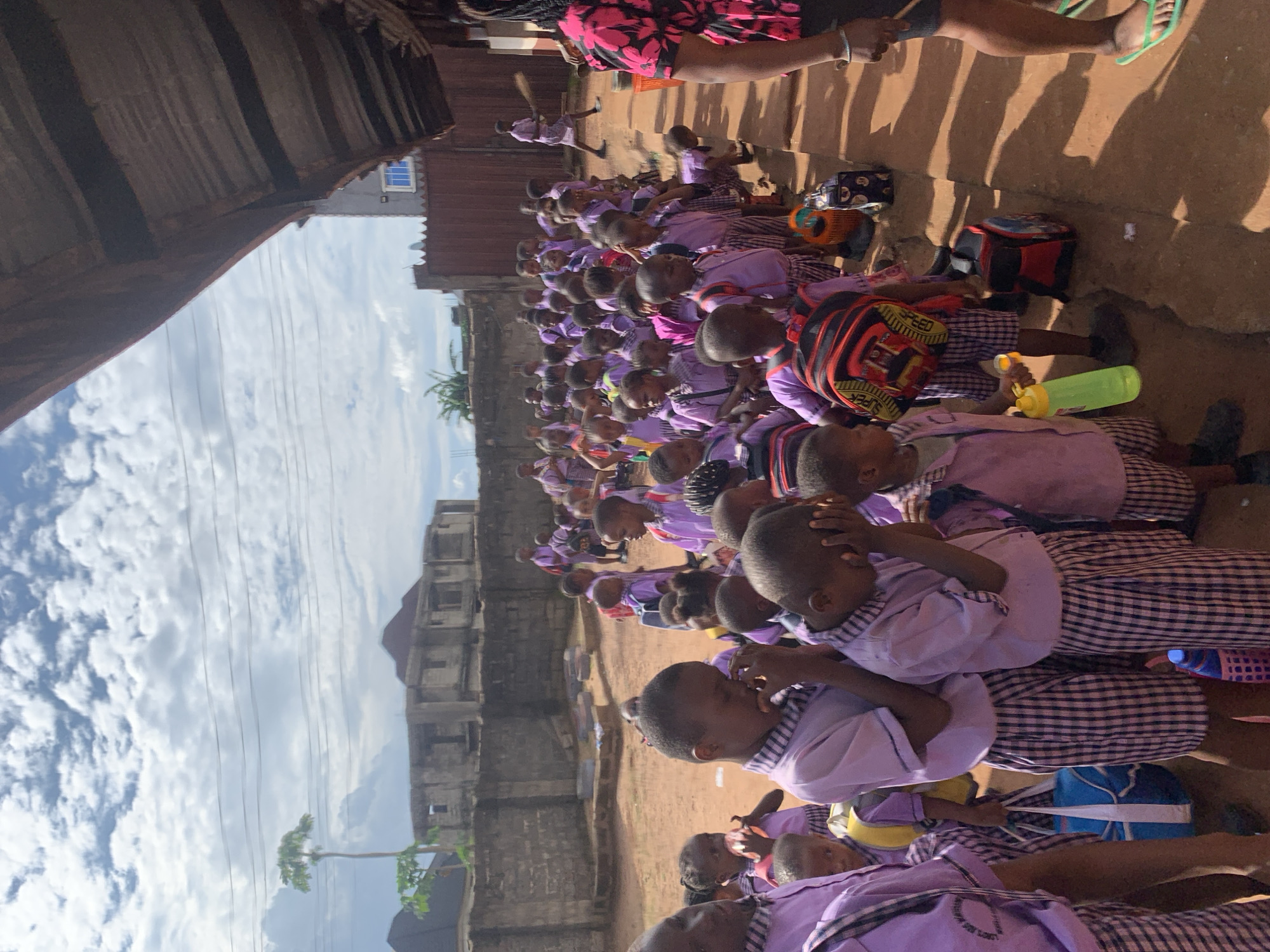 In Ukwu-Abwa, Nigeria, a large group of children in matching purple and checkered uniforms are sitting on the ground in an outdoor area. The children appear to be assembled, possibly in a schoolyard or gathering space adjacent to buildings and a fence. Some children have backpacks and water bottles with them. A few adults are present. Overhead, power lines stretch across a partially cloudy sky. The scene suggests a school or community event in a developing urban area.