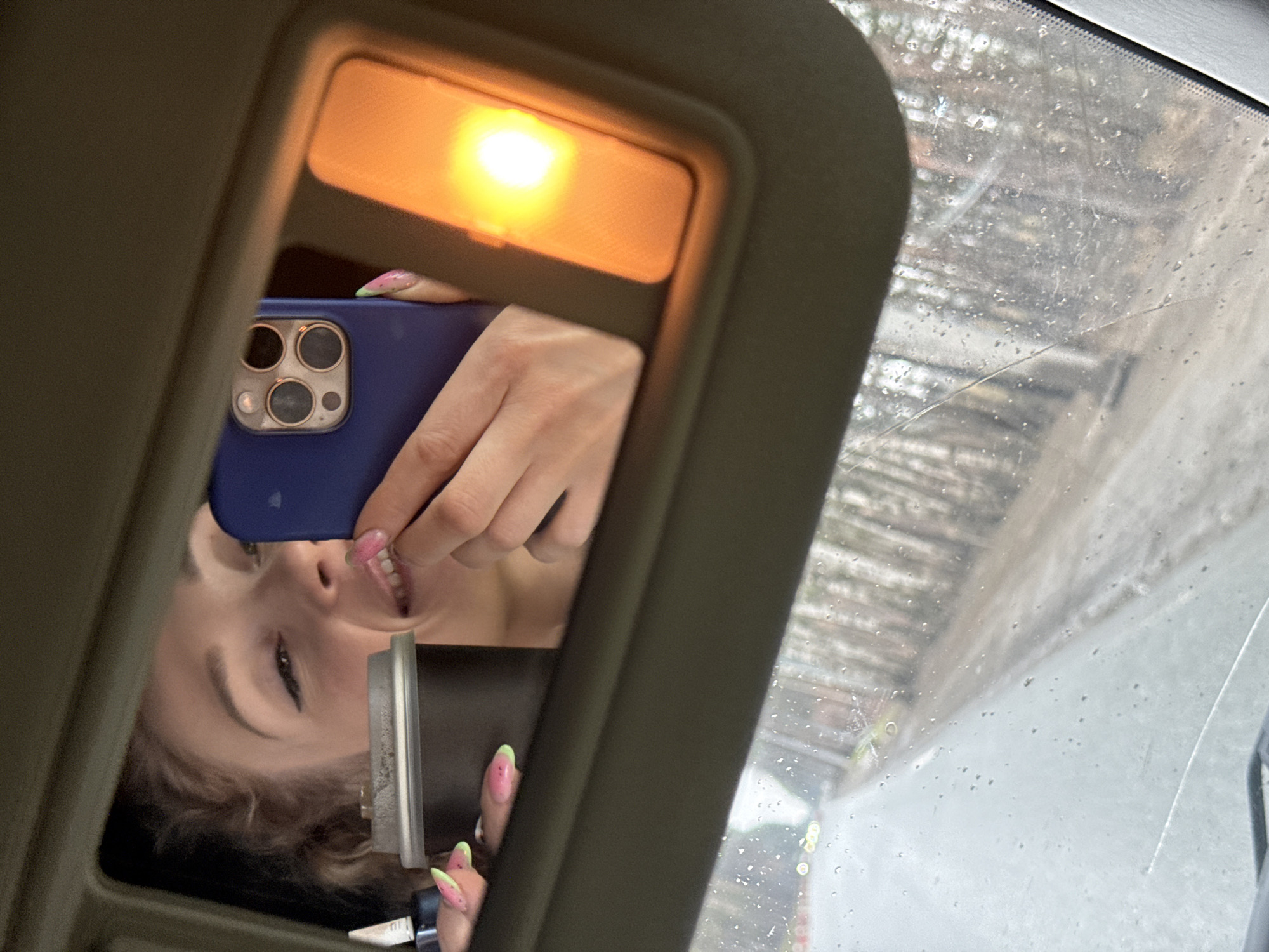 A woman is taking a selfie in the mirror of a car's sun visor. She's holding a blue smartphone, and the mirror is lit by an overhead lamp. Outside the windshield, the scene is wet, possibly raining, and shows an urban street. The location may be Perm, Russia. The windshield has cracks and water droplets.