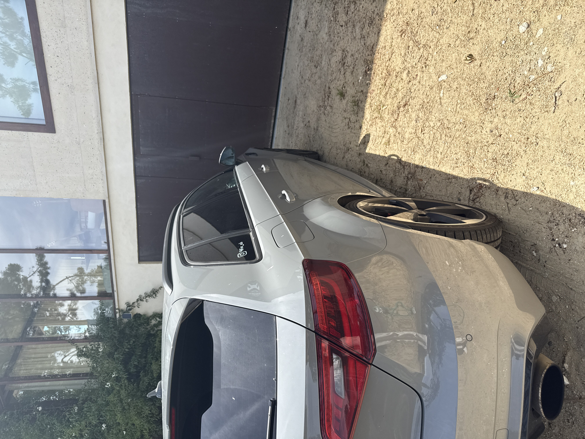 A light grey hatchback car is parked on a sandy or gravelly surface next to a modern building. The image is oriented with the car's right side and rear facing the viewer. The ground is dry, strewn with small stones, sparse weeds, and some debris like pine needles. Strong sunlight creates distinct shadows, indicating a clear, sunny day.
The car features a sleek design with dark-tinted rear windows and a prominent, large black exhaust pipe visible on the right side of the rear bumper. It has black, multi-spoke wheels with low-profile tires. The rear taillights are red, and a circular parking sensor is visible on the light grey bumper. The car appears somewhat dusty. A black roof spoiler is present above the rear window. On the rear side window, a sticker displays the text "@Min_cli" and a small skull-like graphic underneath it.
To the left, a modern building with a light-colored, possibly stone or concrete, facade is visible. It features large, dark-framed windows that reflect tall trees and the sky. Green foliage or climbing plants grow along the base of the building, below the lower windows. A dark-colored wall or garage door is seen further back. The overall scene suggests a private residence in a sunny, possibly coastal or semi-rural environment, consistent with Lège-Cap-Ferret, France. No people or activity are present.