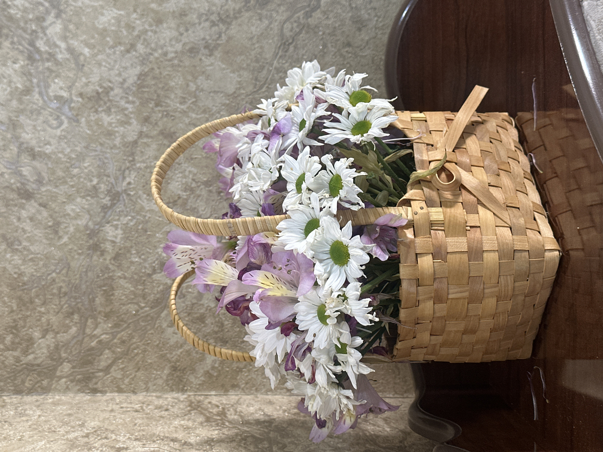 A woven wicker basket filled with a bouquet of white daisies and purple alstroemeria flowers sits on a polished wooden surface. The basket has a curved handle and a small wooden bow tied around one side. The background consists of a textured wall with a pattern resembling marble or stone. The scene is lit from above, casting soft shadows. No people or animals are present. The location is not identifiable as Samarkand City, Uzbekistan, based on the visual information provided.