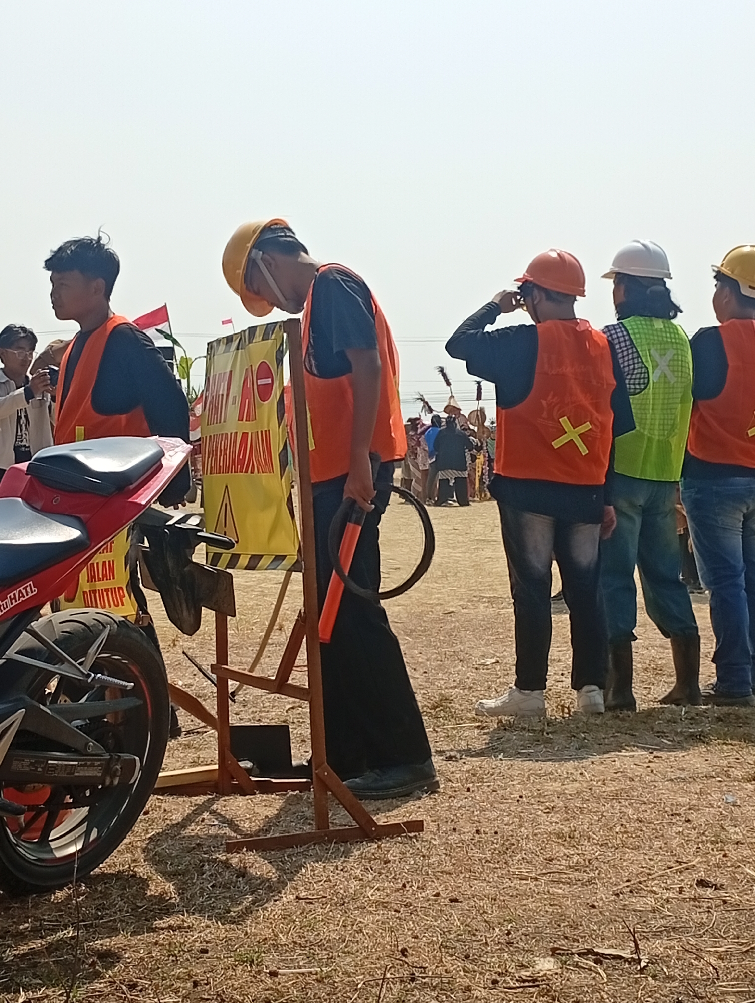 The image depicts a daytime outdoor scene in Bajingjowo, Indonesia. In the foreground, a red motorcycle is partially visible on the left. A group of individuals, primarily wearing orange safety vests and hard hats, are positioned throughout the frame. Some are facing away from the camera, while others are partially turned. One individual, in the center-left, is wearing a yellow hard hat and appears to be looking down, holding an orange object. Behind them, a yellow sign with red text and symbols is propped up. Further into the background, other individuals in traditional attire are visible, suggesting a cultural event or gathering. The ground is covered in dry grass and dirt. The sky is clear and bright.
