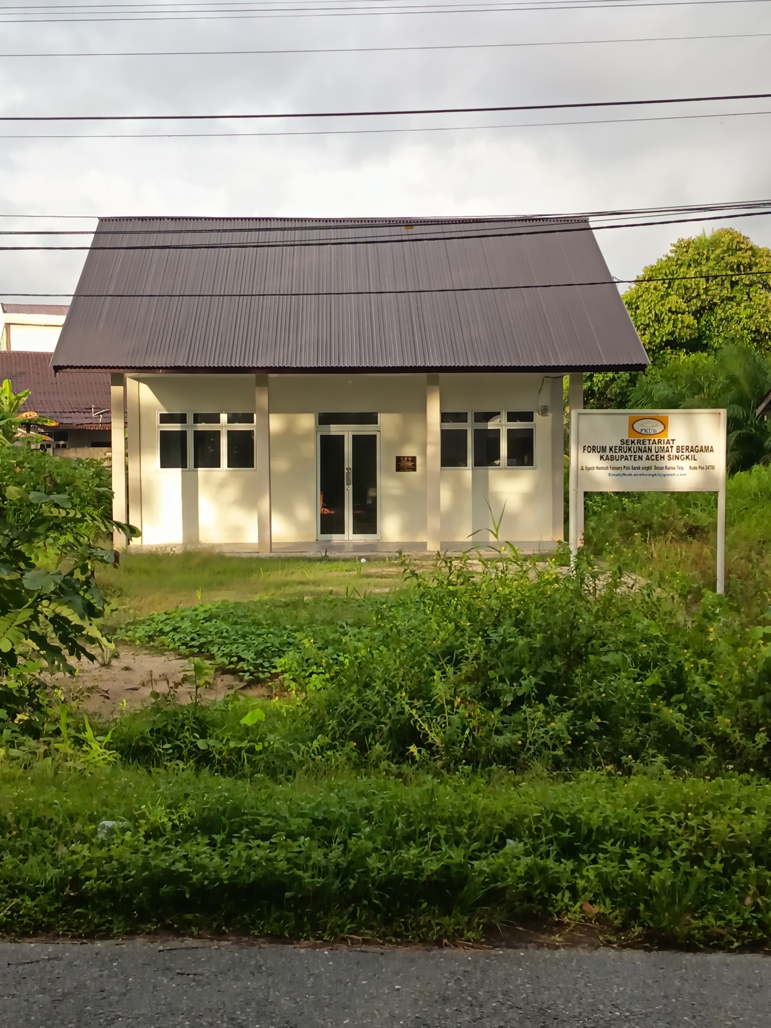 The image shows a small, single-story building with a dark, corrugated metal roof and light-colored walls. The building has a porch supported by pillars and features several windows with white frames and a double glass door entrance. To the right of the building is a white sign with text in Indonesian, which appears to be the official signage for the "Sekretariat Forum Kerukunan Umat Beragama Kabupaten Aceh Singkil" (Secretariat of the Religious Harmony Forum for the Regency of Aceh Singkil). The sign includes an address: "Jl. Syekh Hamzah Fansury Polo Baruh Singkil, Dusun Kuranjo Teip, Kode Pos 24785" and an email address: "fkub.acehsingkil@gmail.com". The building and sign are surrounded by lush green vegetation, including trees and ground cover, suggesting a semi-rural or suburban environment. The sky is overcast with power lines visible across the top of the frame.