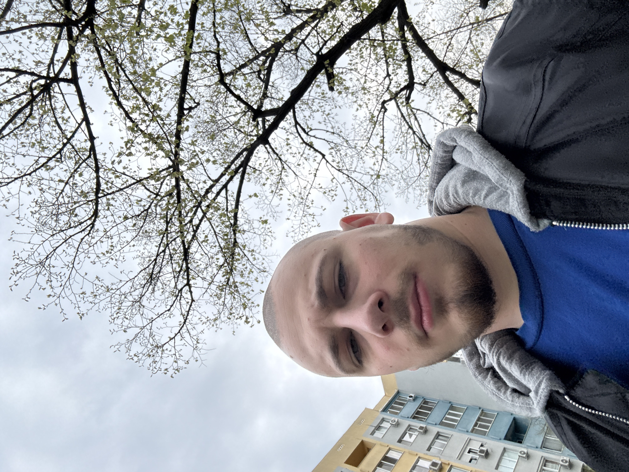 A man is the subject of this photograph, taken from a low angle. He has fair skin, a shaved head, and a short beard. He is wearing a blue shirt under a black jacket with a grey hood. In the background are bare tree branches with small leaves, a cloudy sky, and a building with multiple windows and air conditioning units. The building is painted in shades of yellow and blue. The location may be in Stara Zagora, Bulgaria. The man appears to be taking a selfie.