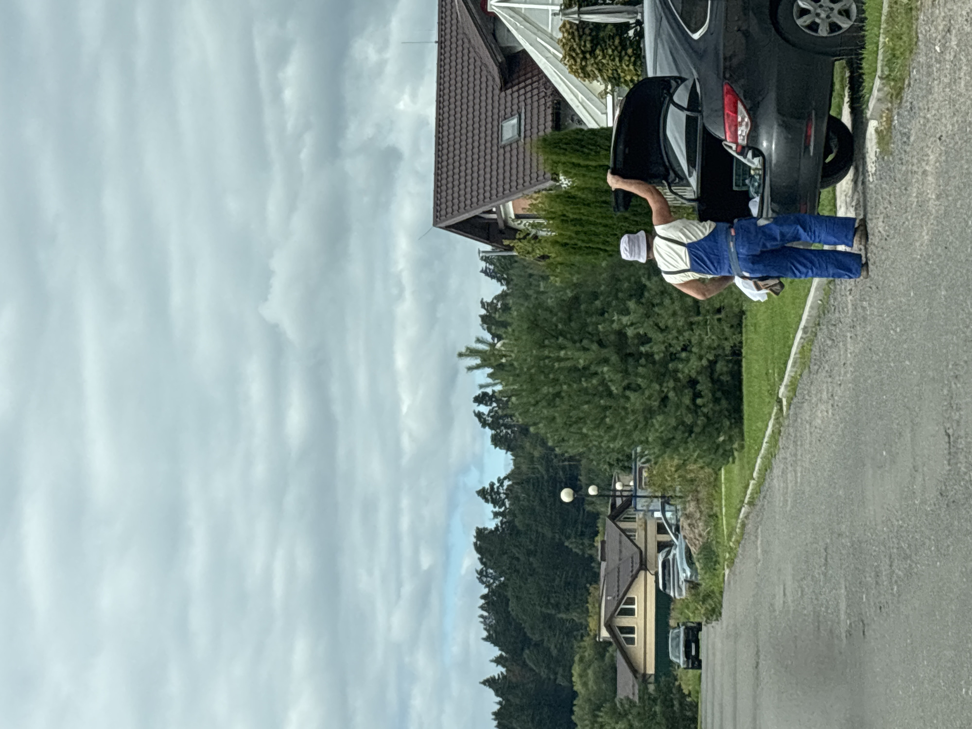 A person wearing blue overalls and a white bucket hat is standing next to the open trunk of a dark-colored car, placing items into it. The car is parked on the side of a paved road. In the background, there are houses with brown roofs, green trees, and a cloudy sky. The location is Zvenigorod, Russia, a town known for its scenic natural surroundings and historical sites.