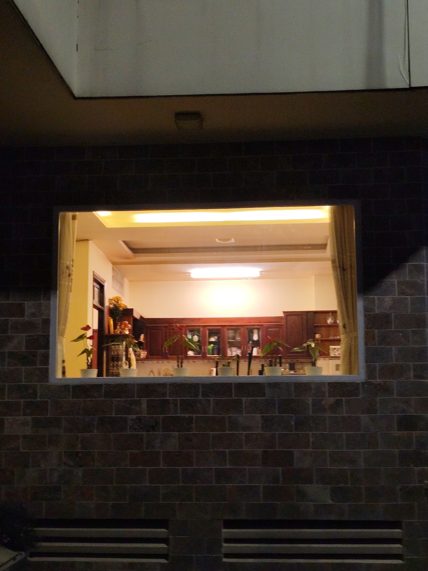 A window in a brick wall reveals an interior space, likely a kitchen, illuminated by ceiling lights. The kitchen features dark wood cabinetry and a counter populated with potted plants and objects. Through the window, hints of a yellow wall and interior design elements are visible. The exterior is dark, with the brick wall suggesting the building is possibly located in Da Lat, Vietnam. The wall and ceiling are black around the white square window.
