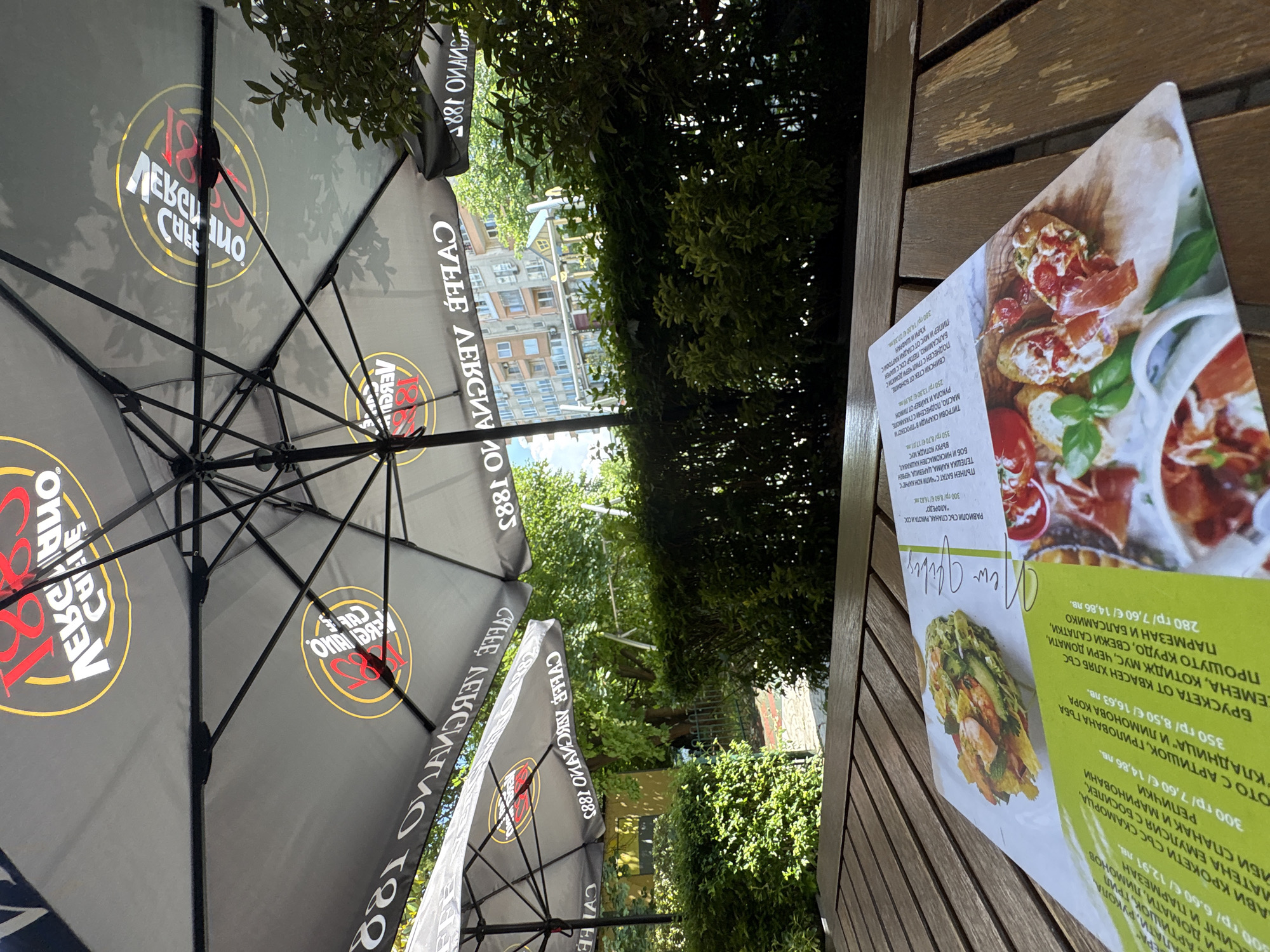 An outdoor cafe scene, rotated 90 degrees clockwise, captures a bright, sunny day in what appears to be Burgas, Bulgaria. The foreground is dominated by the underside of several large, dark grey patio umbrellas, featuring the brand "COFFEE АЛЕКСИНО 1885" in white, red, and gold lettering. The intricate black metal framework of the umbrellas is clearly visible.
To the right, a wooden slatted table holds a printed menu card, open to display various food items with accompanying images. The menu, written in Bulgarian, lists appetizers, salads, and main courses with prices in both Euros (€) and Bulgarian Levs (лв.). Notable items include "БРУСКЕТА ОТ КВАСЕН ХЛЯБ СЪС СЕМЕНА, КОТИДЖ МУС, ЧЕРИ ДОМАТИ, ПРОШУТО КРУДО, СВЕЖИ САЛАТКИ, ПАРМЕЗАН И БАЛСАМИКО" (bruschetta with prosciutto crudo, parmesan, and balsamic), "ТИГРОВИ СКАРИДИ В "ПРОСЕКО"" (tiger prawns in Prosecco), "РАВИОЛИ СЪС СПАНАК, РИКОТА И СОС "АЛФРЕДО"" (ravioli with spinach, ricotta, and Alfredo sauce), and "СВИНСКИ СТЕК ОТ БОНФИЛЕ" (pork tenderloin steak). Prices range from around 6.60€/12.91лв for salads to 13.80€/26.99лв for prawns.
Beyond the umbrellas, lush green trees and foliage provide a natural backdrop, with glimpses of a multi-story building with windows in the distance, reflecting the bright sky. The overall atmosphere suggests a relaxing outdoor dining experience on a clear day.
