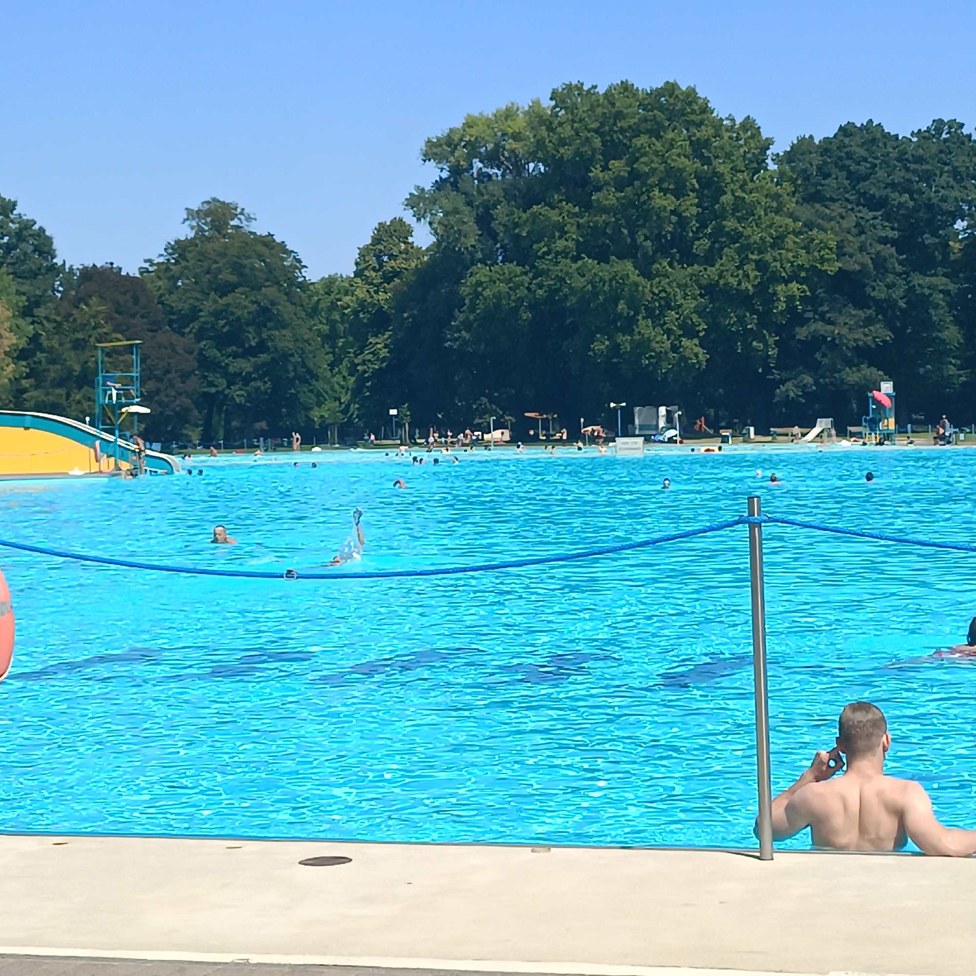A wide-angle, eye-level outdoor photograph captures a sunny day at the Opelbad swimming pool in Frankfurt, Germany. The bright blue water of the pool dominates the foreground, with the edge of the pool visible in the bottom center of the frame. A man with short brown hair is partially submerged in the water, facing away from the camera, holding a mobile phone to his right ear with his right hand, and resting his left arm on the pool edge. In the mid-ground, several other individuals are in the pool, with some swimming and others appearing to be relaxing. To the left, a yellow and blue water slide structure with a lifeguard tower is visible. The background consists of a dense line of mature green trees, with a clear blue sky above.