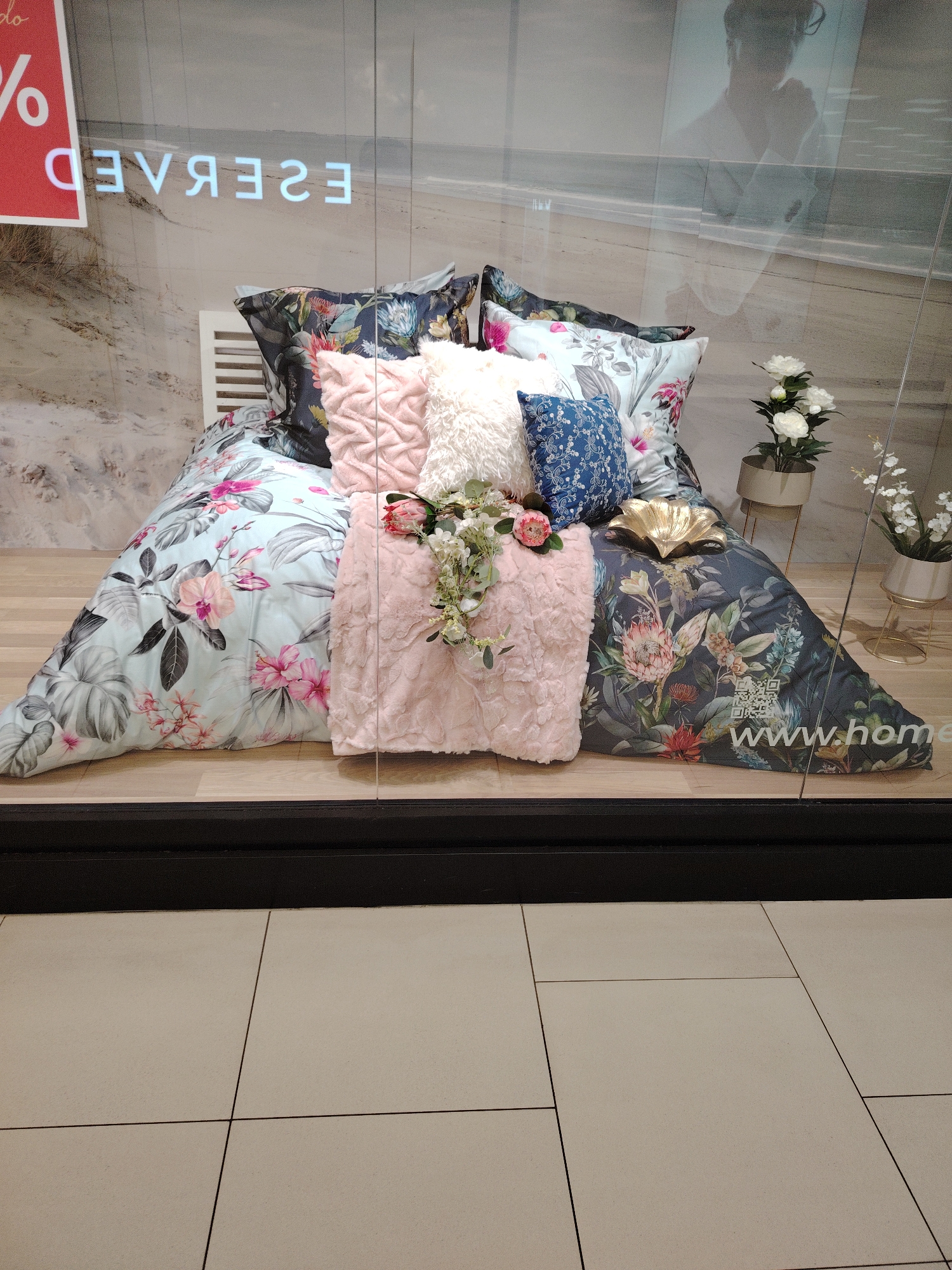 The image shows a retail display of bedding in a store window. The display features a bed with floral-patterned duvet covers and pillows, including a pink textured pillow and a blue patterned pillow. There are also decorative items such as a gold shell-shaped object and artificial flowers. In the background, a blurred image of a beach and a silhouette of a person are visible, likely reflections on the glass. Text on the window includes "RESERVED" and "www.home" along with a QR code. The display is set on a wooden floor, and the foreground shows beige tiled flooring. The location of this display is not specified within the image or its context.
